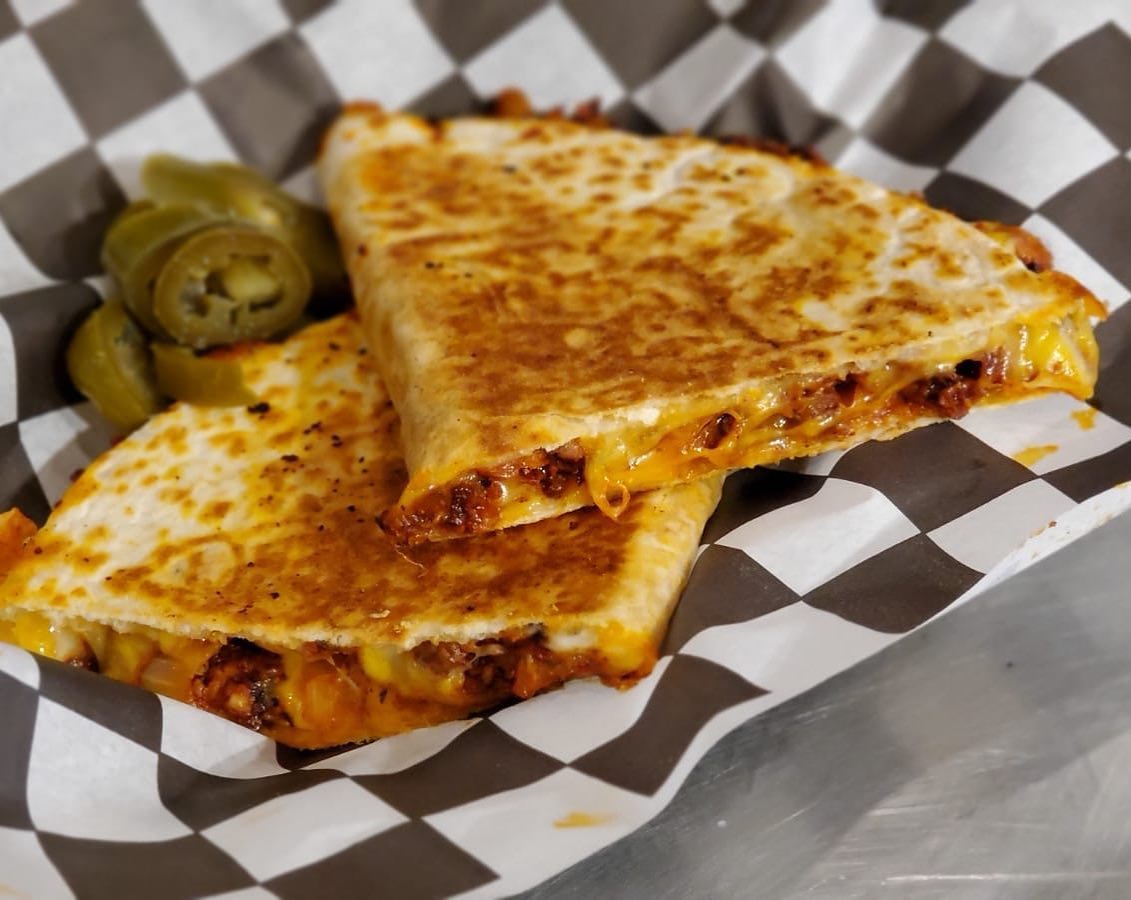 A close-up of a quesadilla cut into triangles, filled with melted cheese and meat, served on a black and white checkered paper with a side of sliced jalapeños.