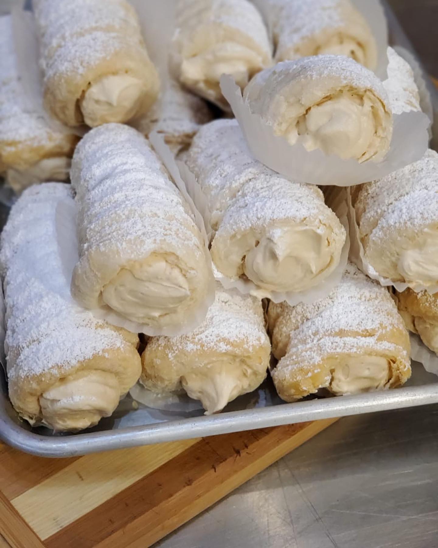 A tray of freshly made cannoli dusted with powdered sugar, displayed with creamy fillings visible at each end. the pastry shells have a golden-brown color and flaky texture.