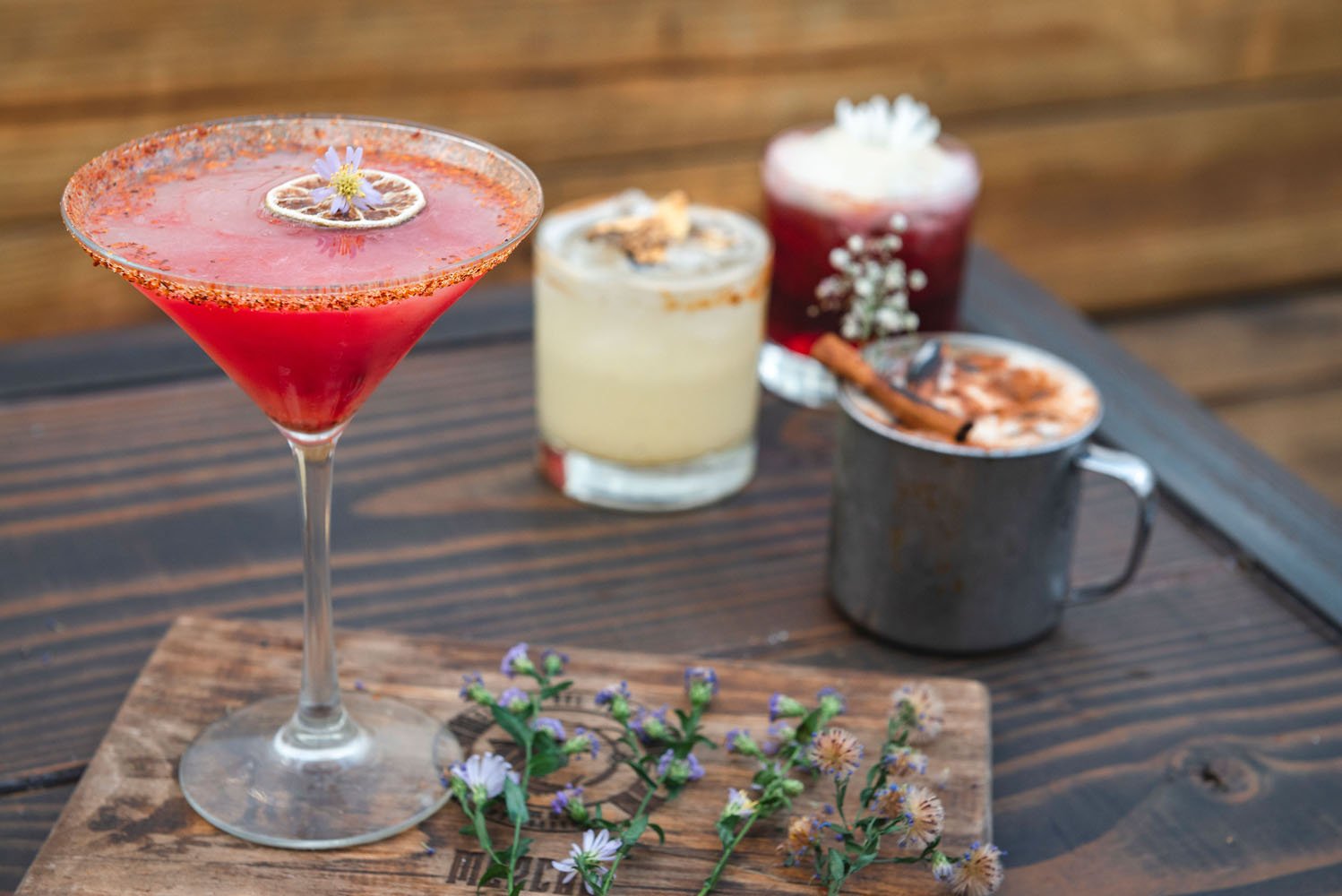 A variety of four cocktails on a wooden table, including a margarita, a moscow mule, and berry-flavored drinks, garnished with flowers and spices.