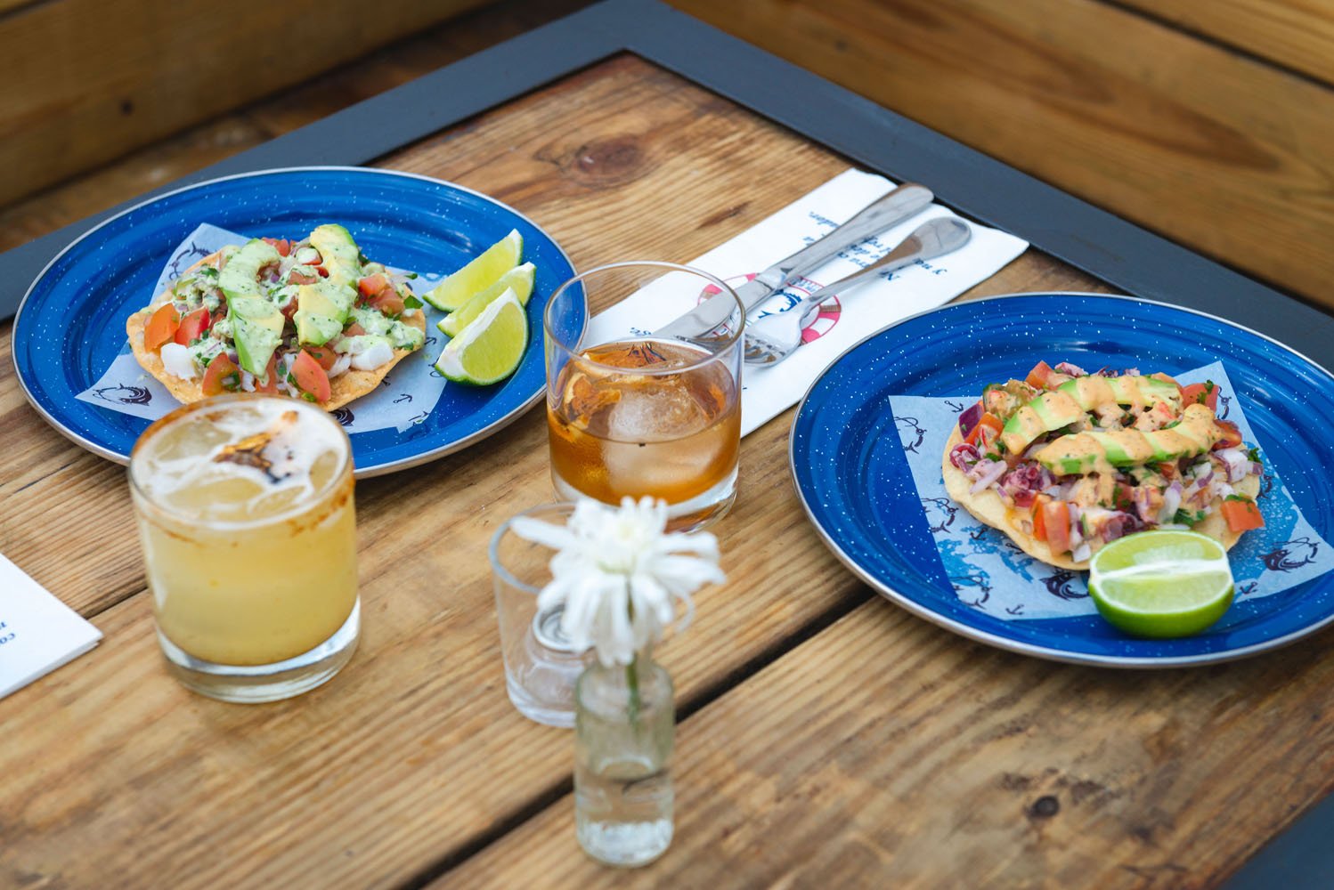 Two blue plates each containing colorful fish tacos, paired with glasses of iced tea, on a wooden table with cutlery and a small white flower vase.