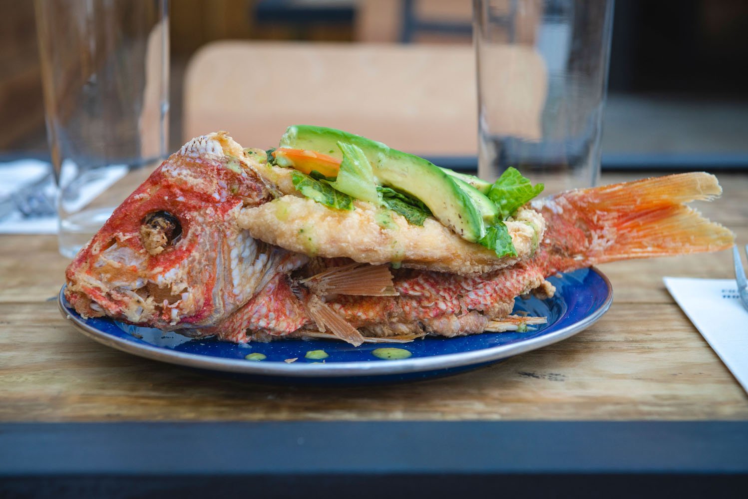 A plate with a whole red snapper, garnished with green vegetables, at a restaurant table, with a glass of water and background seating visible.