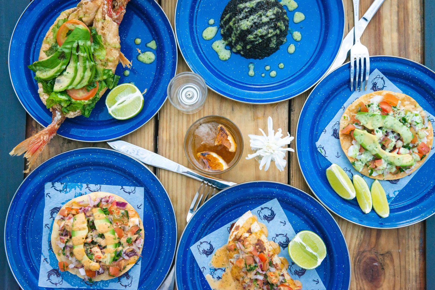 A variety of mexican dishes on blue plates, including shrimp tacos and enchiladas, spread on a wooden table, garnished with lime wedges.