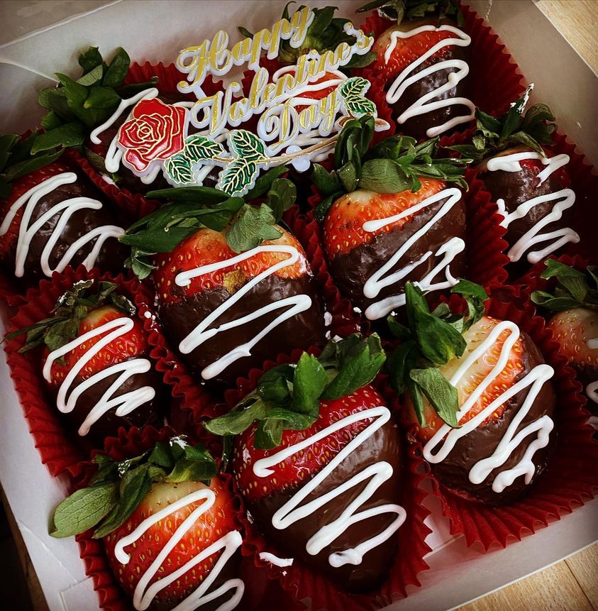 A box of chocolate-dipped strawberries with decorative white and orange icing, accompanied by a "happy valentine's day" message and a rose, all displayed in red cupcake liners.