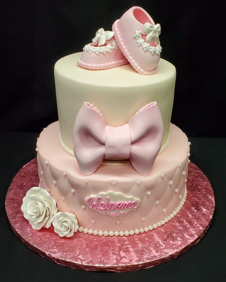 A two-tier baby shower cake, decorated in pink and cream colors, features a large pink bow, white roses, and tiny baby shoes on top; the lower tier is adorned with the name "valeria" in cursive script.
