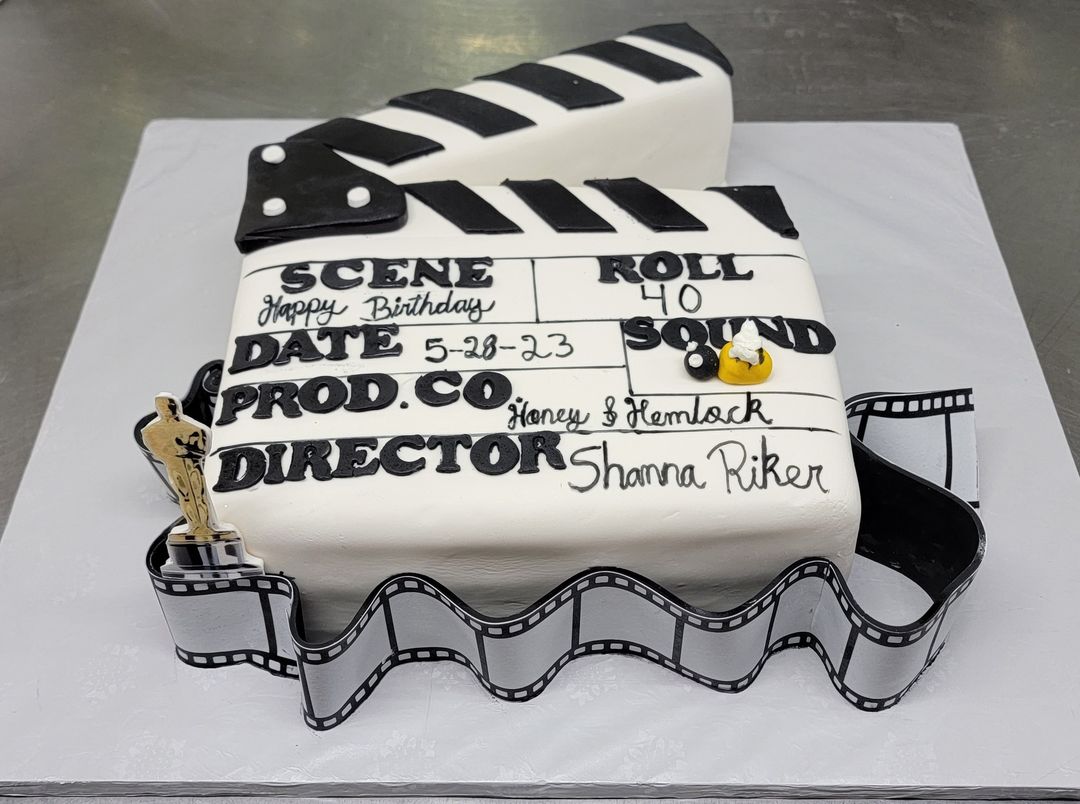 A rectangular birthday cake designed to look like a film clapperboard, with black and white detailing and a film reel decoration, celebrating a birthday for shanna riker.