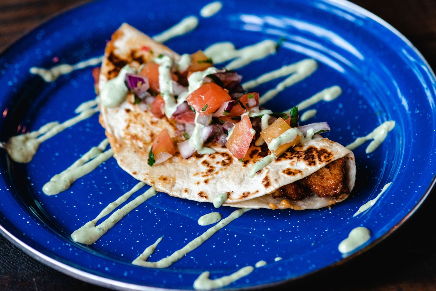 A taco with crispy filling, topped with diced tomatoes, onions, and cilantro, drizzled with a creamy sauce on a vibrant blue plate.