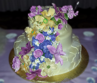 A three-tiered wedding cake adorned with a colorful assortment of edible flowers in shades of purple, blue, and yellow, enhanced by delicate butterflies.