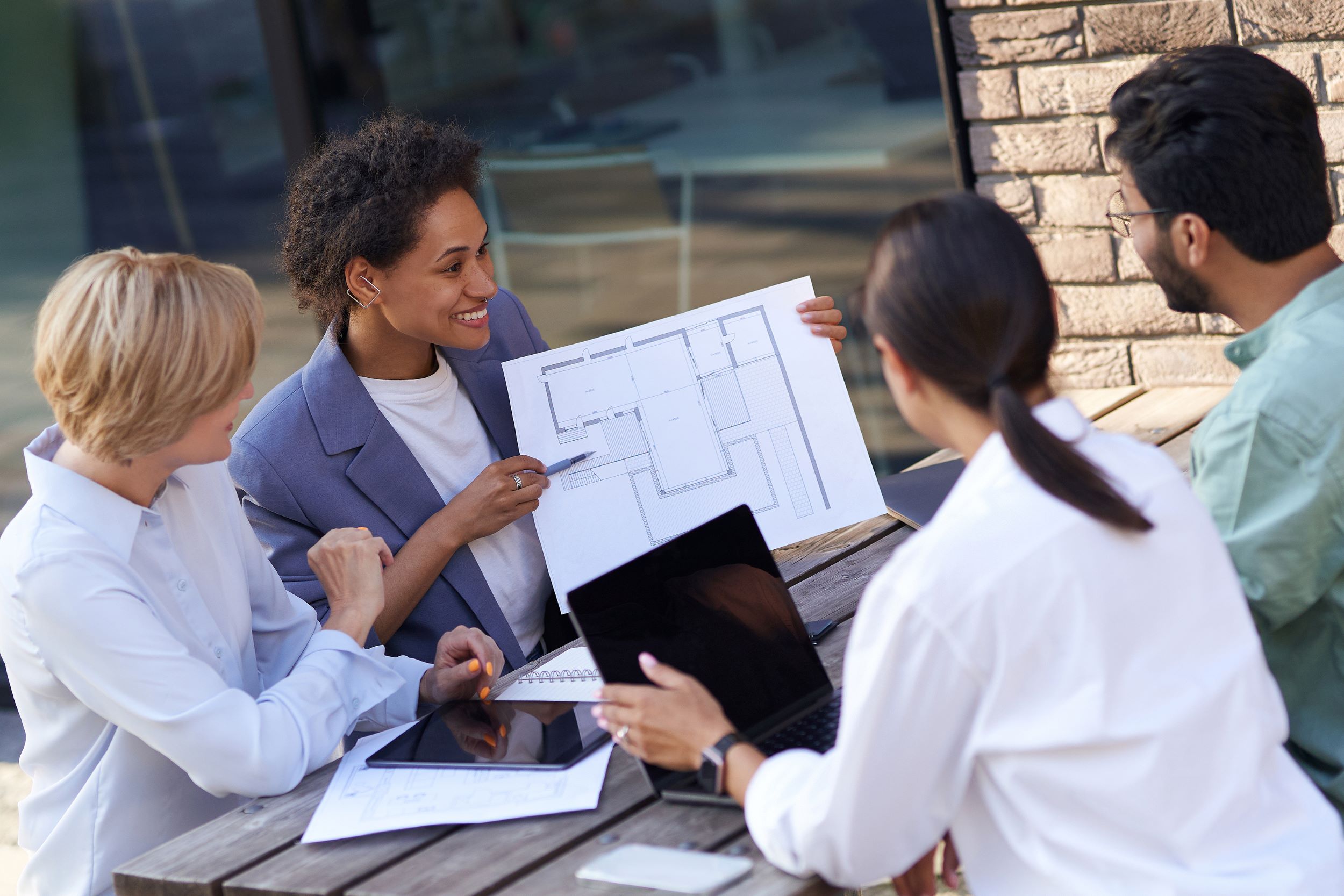 A diverse group of professional colleagues review building plans outdoors, focused on a document held by an african american woman, with a laptop and additional papers on the table.