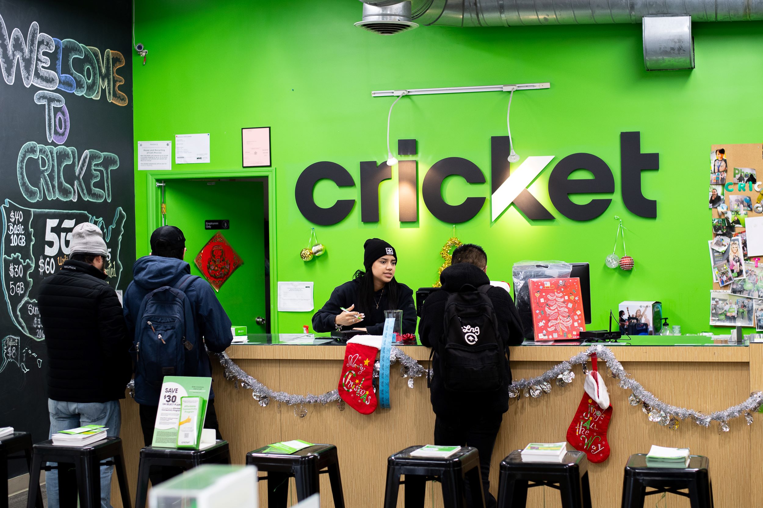 Customers interact with an employee at a cricket store counter, decorated with holiday ornaments and stockings, under a bright green wall with the cricket logo.
