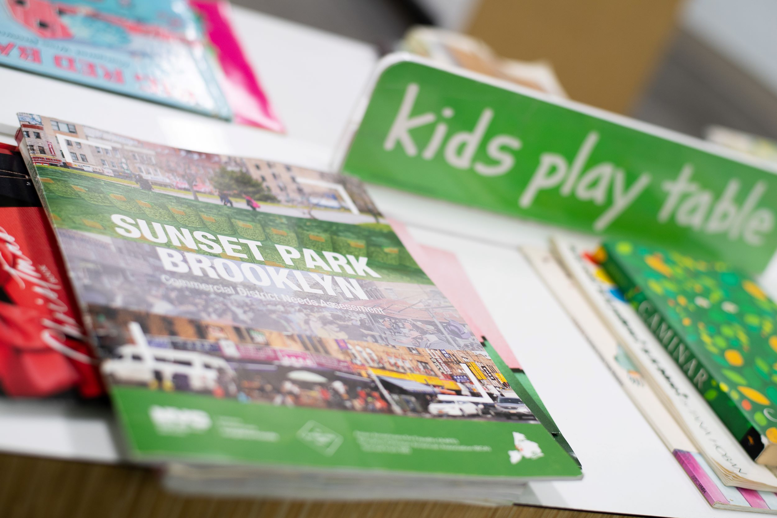 Close-up view of a book titled "sunset park brooklyn" on a table with other books, and a green sign saying "kids play table" in the background, focused on the book cover.