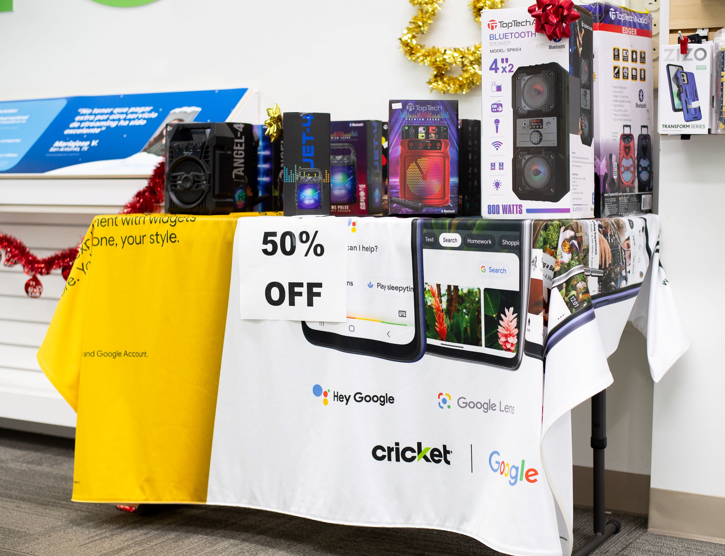 A display table at a cricket store featuring electronic gadgets, such as headphones and speakers, and promotional signs for a 50% off sale, adorned with google branding and holiday decorations.