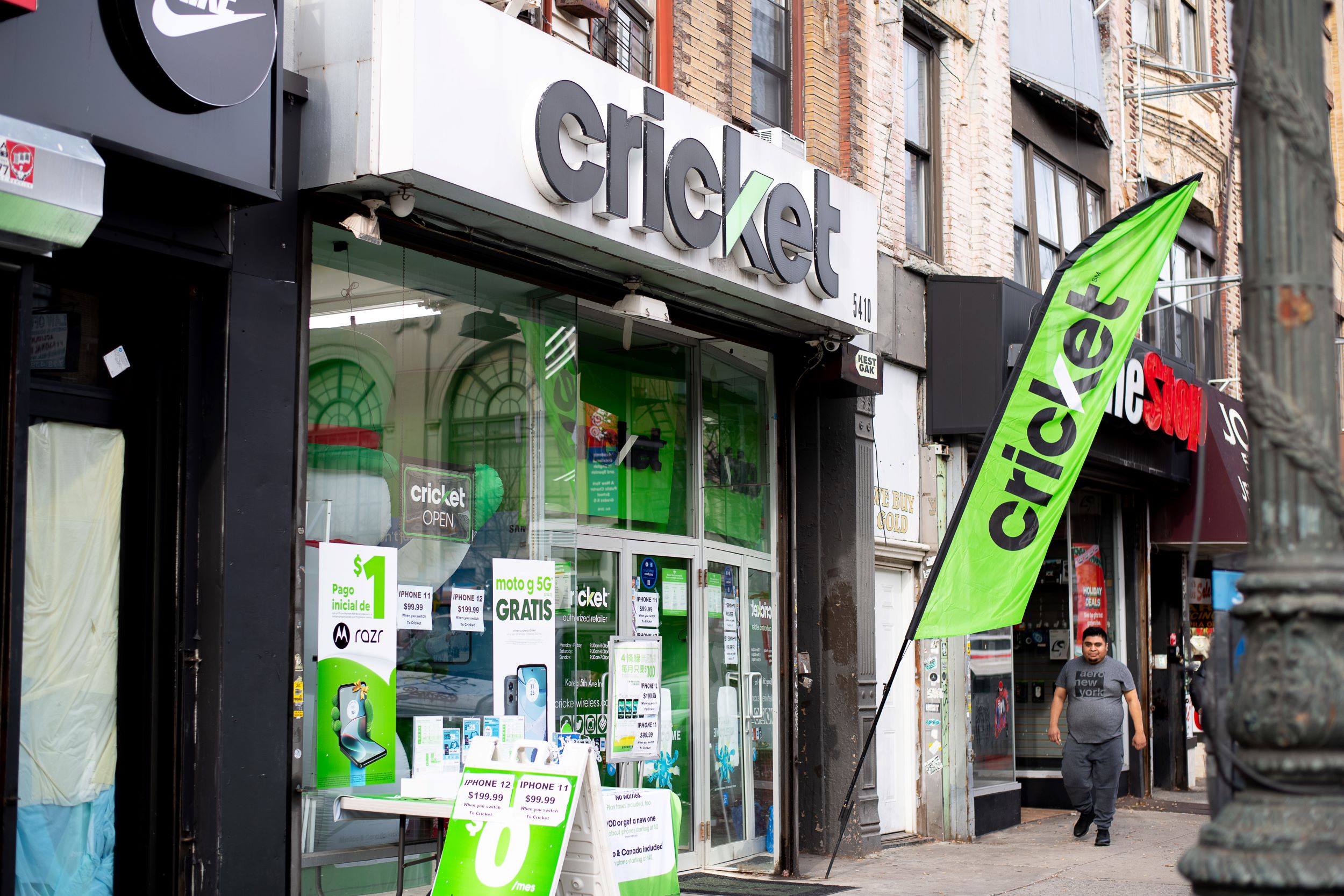 A cricket wireless store on a busy street with a prominent store sign, green promotional flags, and banners advertising deals. a person walks by the storefront.