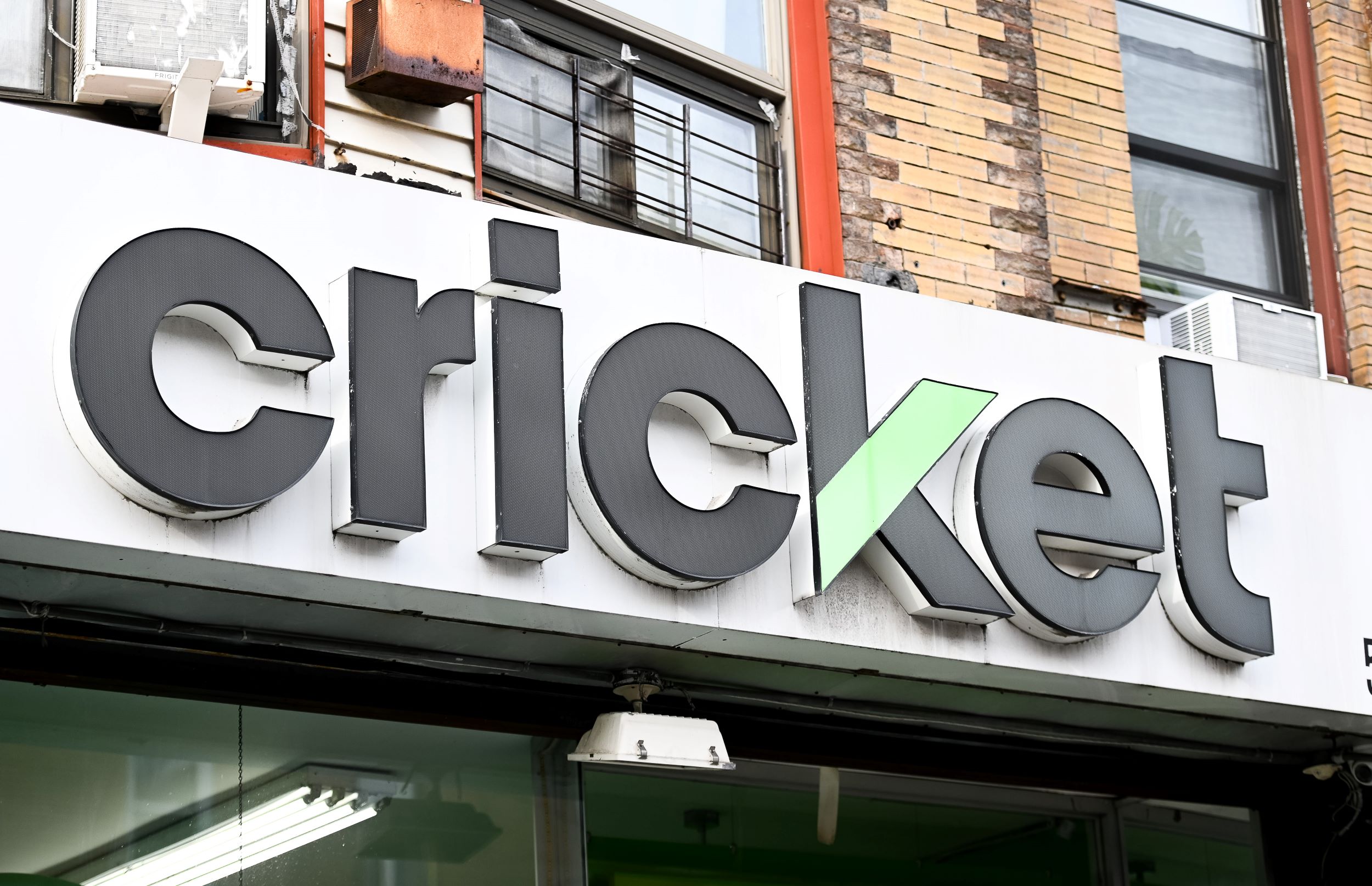 Signage with the word "cricket" in bold, three-dimensional letters mounted on a store facade, featuring large windows and part of an air conditioner unit visible.