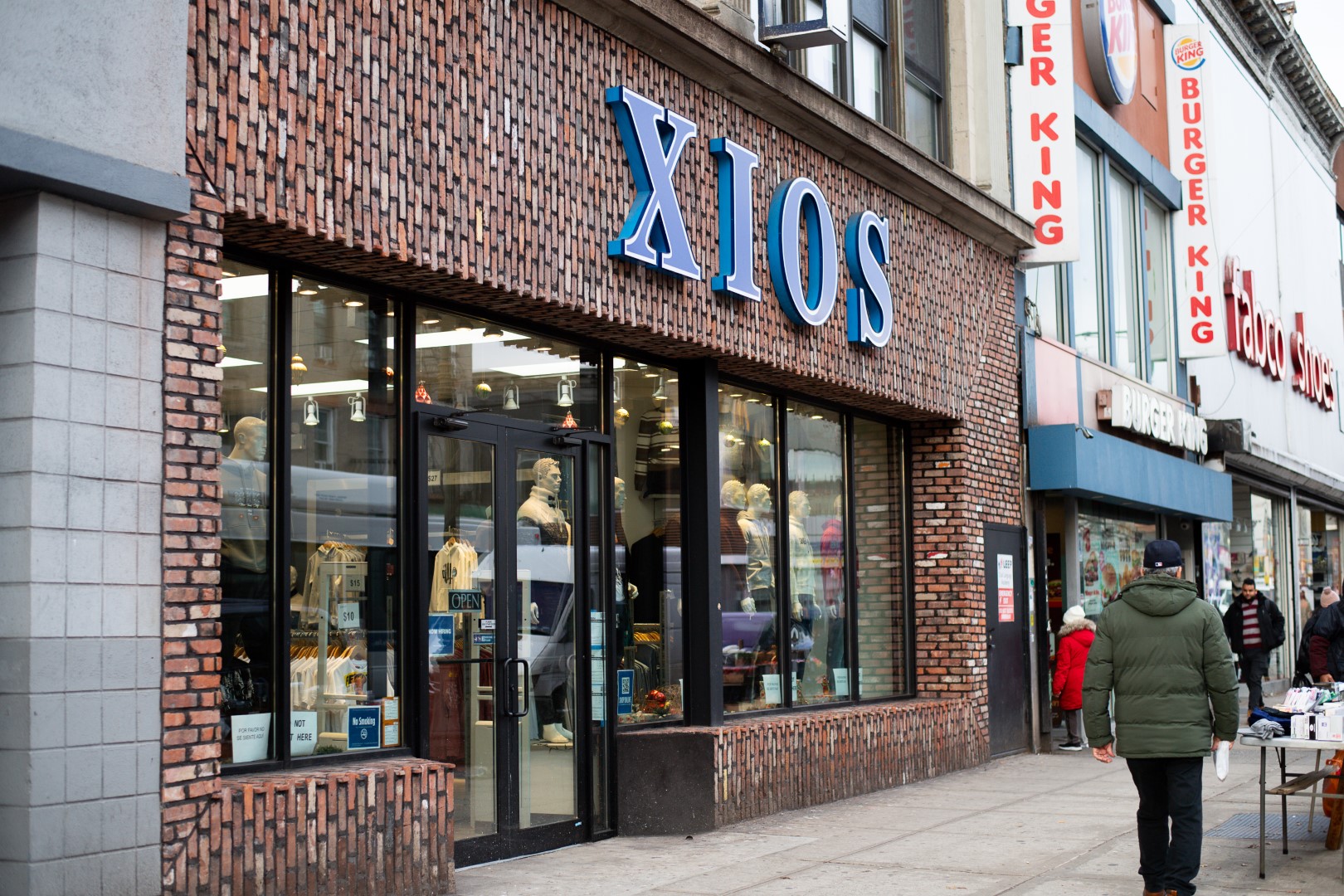 Exterior view of a brick building with a blue sign reading "xios" next to a burger king, featuring large glass windows displaying clothing and a pedestrian walking by.