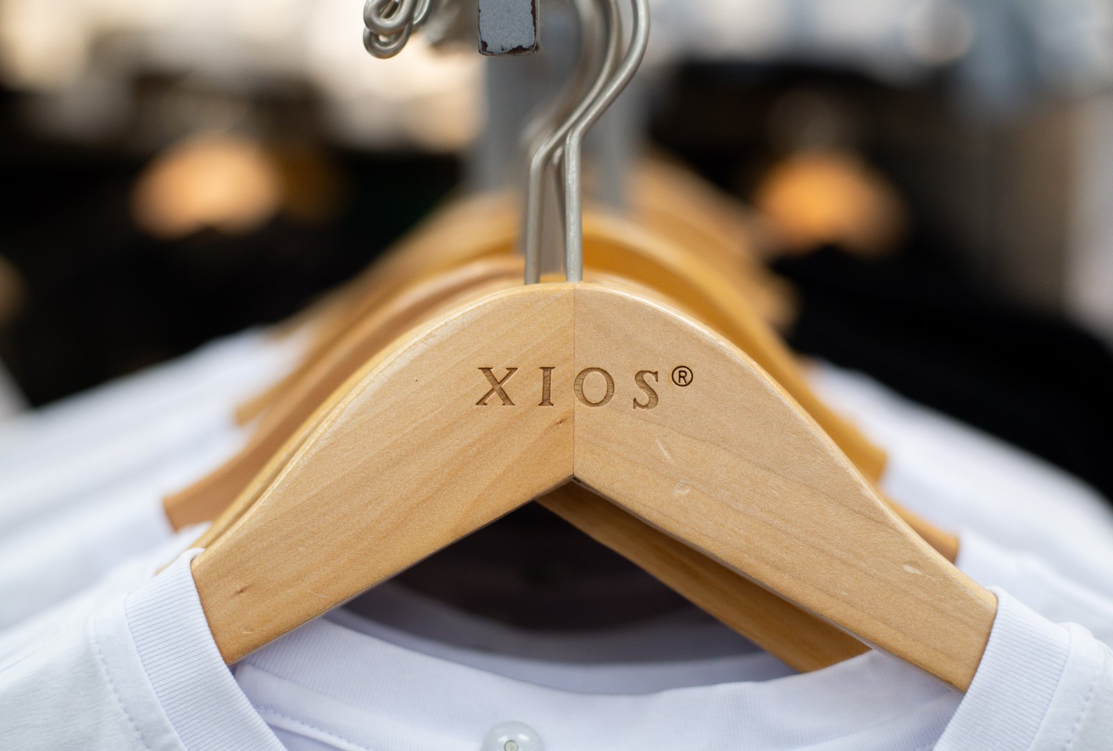 Close-up of a wooden hanger with the logo "xios" engraved, holding white shirts in focus, with a blurry background of additional clothes.