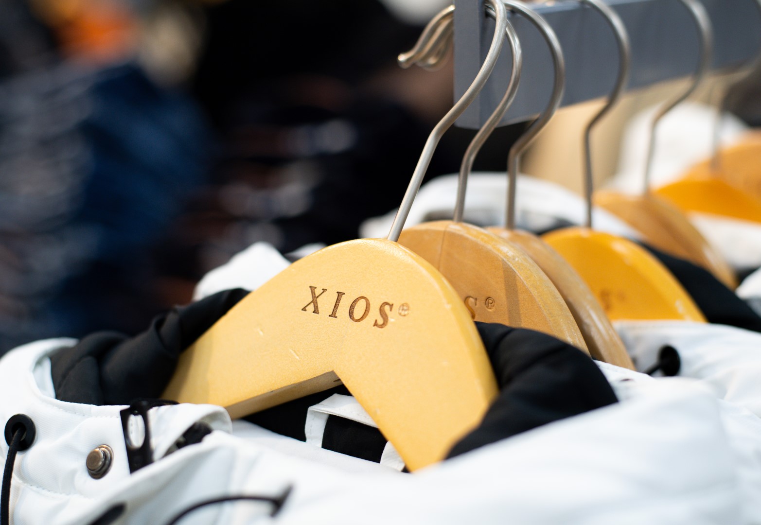 Close-up of white shirts hanging on wooden hangers labeled "xios" in a clothing store, with a blurred background of other apparel.