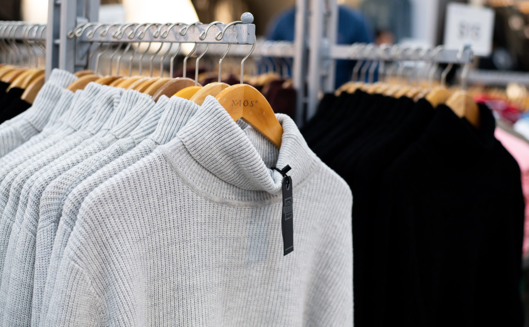 A row of sweaters on hangers in a clothing store, featuring a prominent textured gray sweater in the foreground and various darker clothes in the background.
