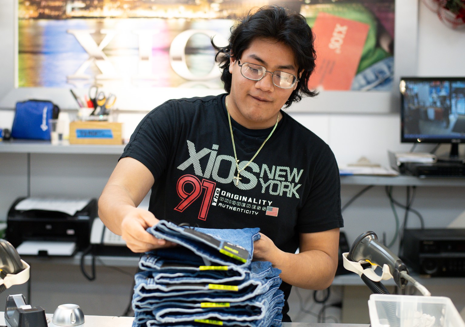 Young man in a black t-shirt stands in a workspace, holding a stack of folded jeans, with design tools and posters visible in the background.