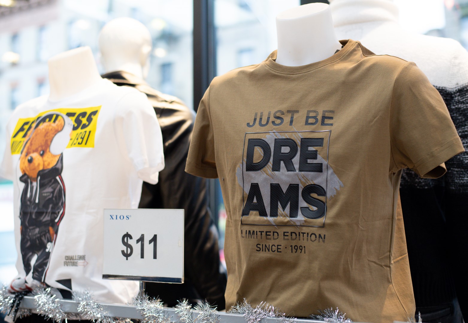 Mannequin displaying a limited edition "just be dreams" t-shirt, with other shirts visible, positioned in a store window adorned with silver festive decoration, price tag showing $11.