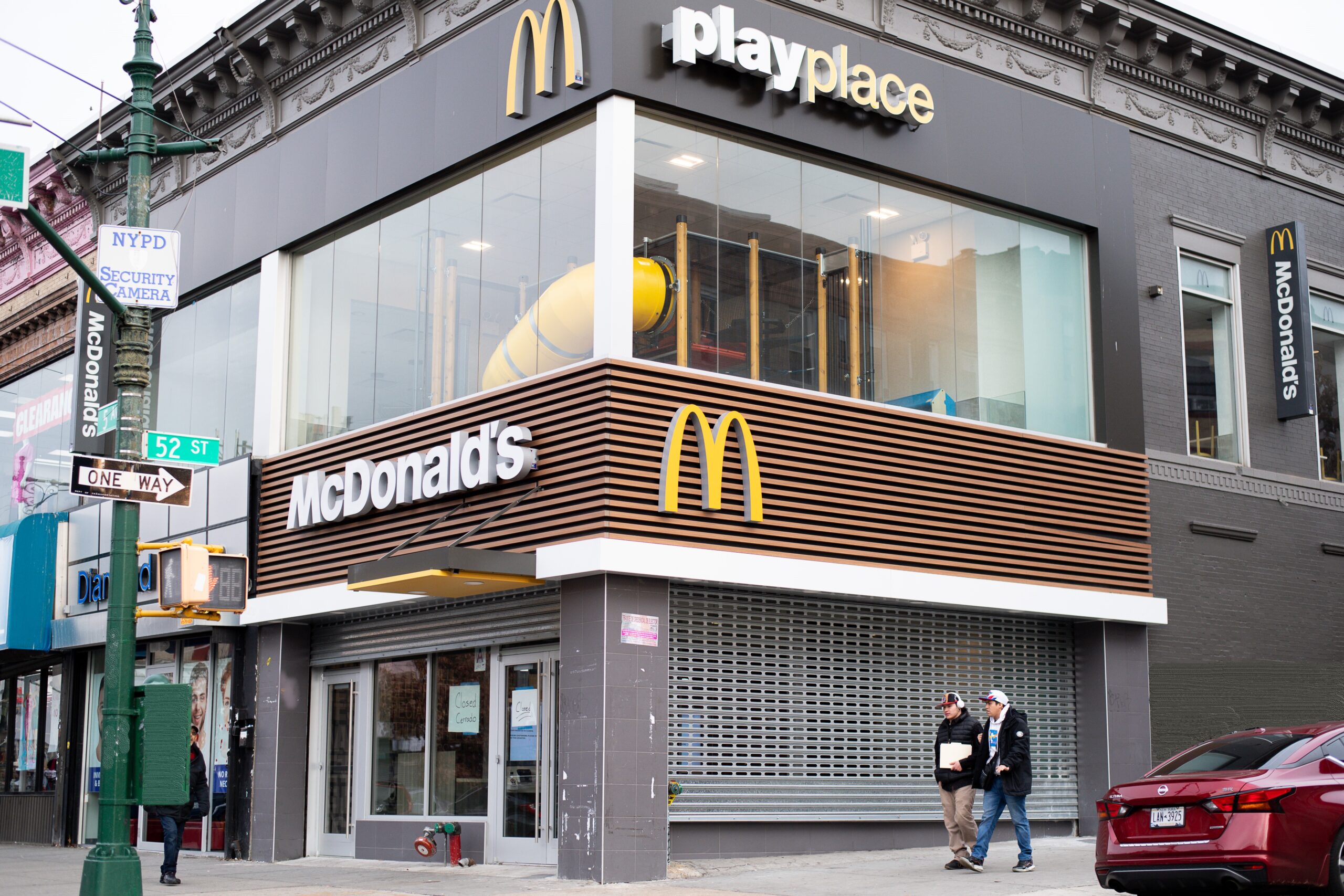 Modern two-story mcdonald's restaurant with playplace, located on a city corner with pedestrians passing by. the facade features large glass windows and distinctive branding.