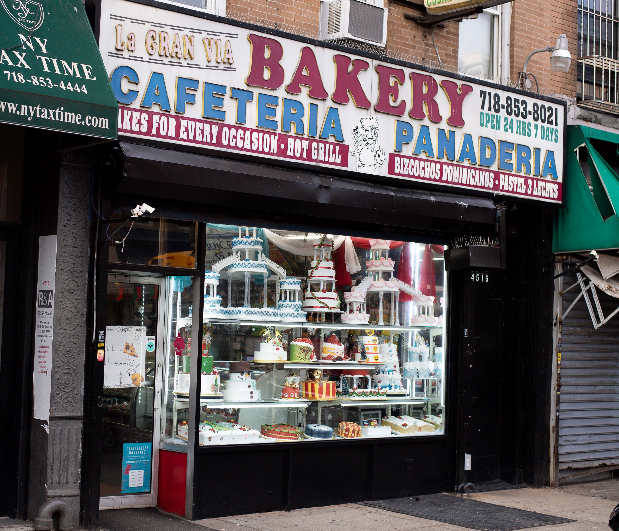 A bakery storefront with a large glass window displaying elaborate cakes. above the window, signs advertise various services in english and spanish, including a cafeteria and bakery items.