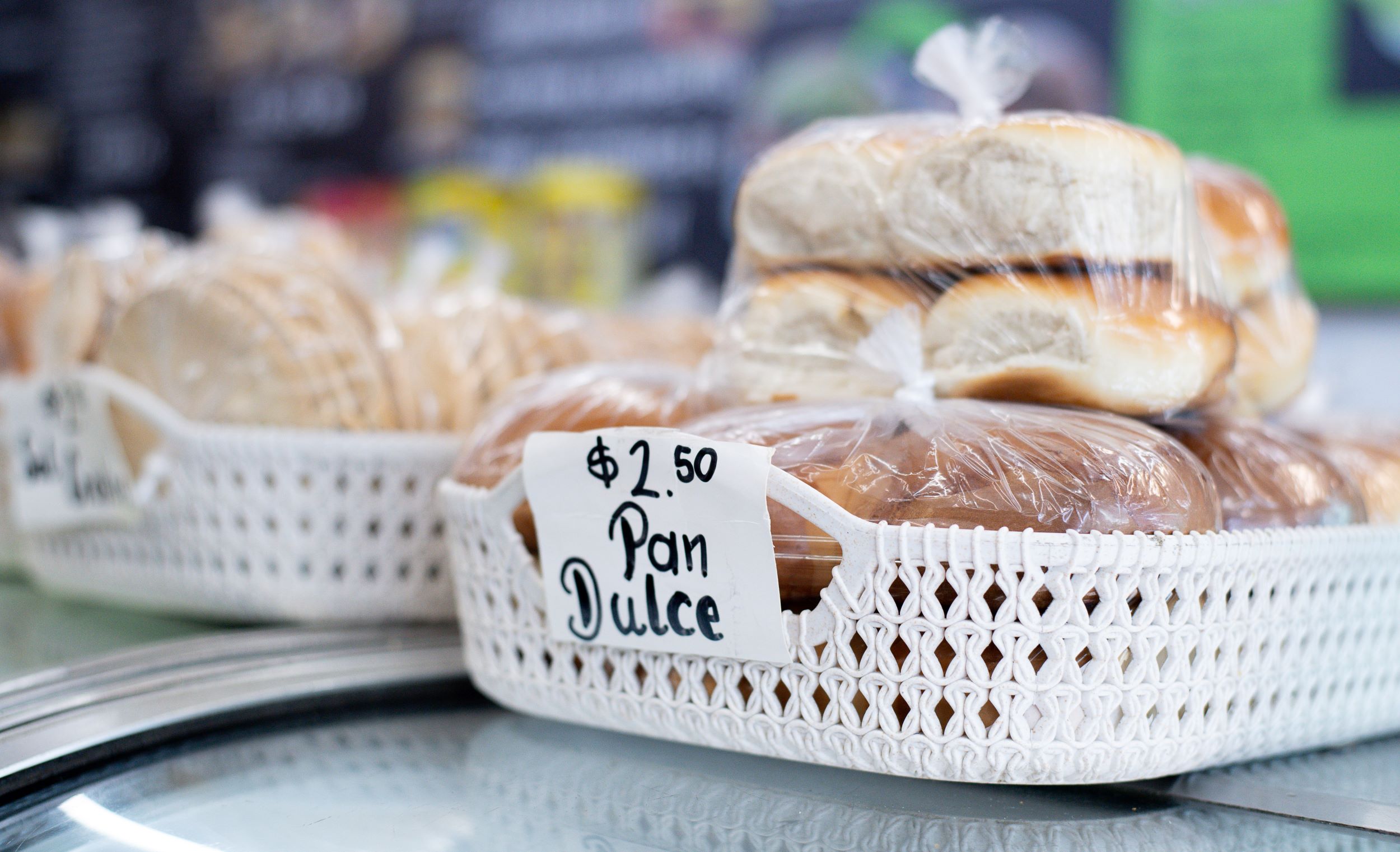 Woven baskets filled with bagged loaves of pan dulce (sweet bread), each priced at $2.50, displayed on a shiny counter in a bakery.