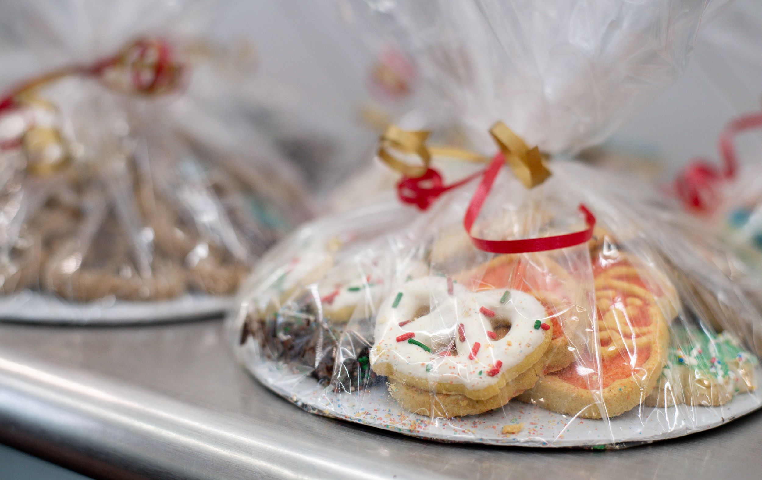 Assorted holiday cookies decorated with sprinkles and icing, packaged in clear plastic bags tied with red and gold ribbons on a metal tray.