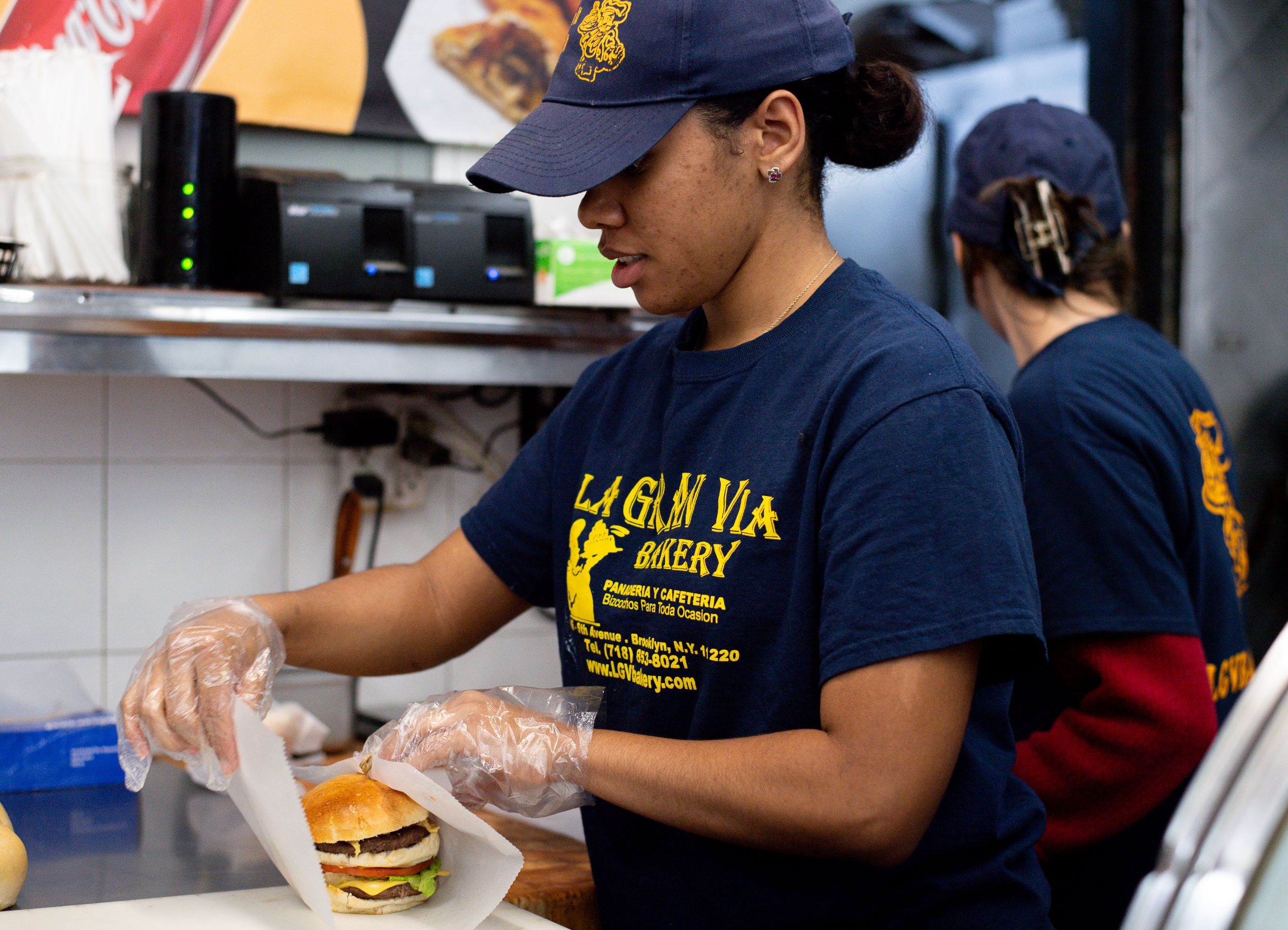A woman wearing gloves and a branded t-shirt prepares a burger in a bustling kitchen, with another worker visible in the background.