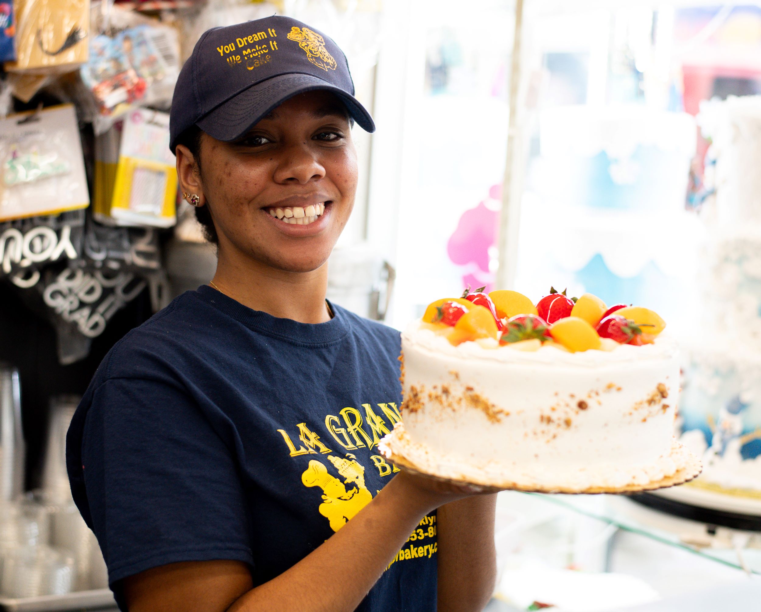 A cheerful bakery employee in a navy cap and t-shirt, presenting a large, beautifully decorated cake with fruit toppings at a colorful dessert shop.