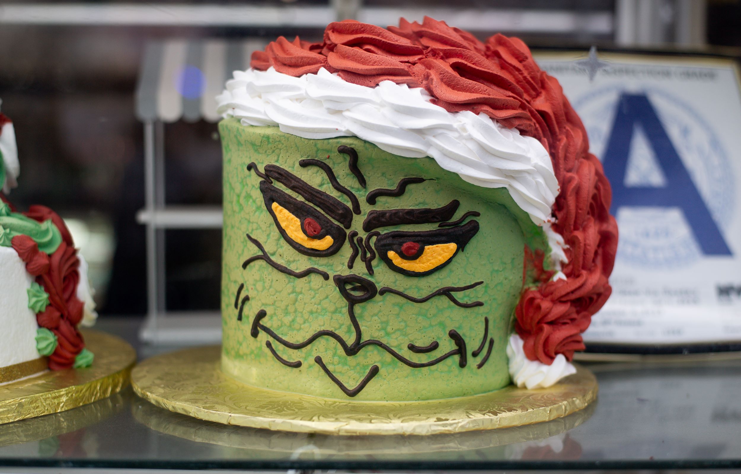 A detailed cake decorated like a green monster face with expressive eyes and a menacing expression, topped with red and white icing, displayed on a gold plate.