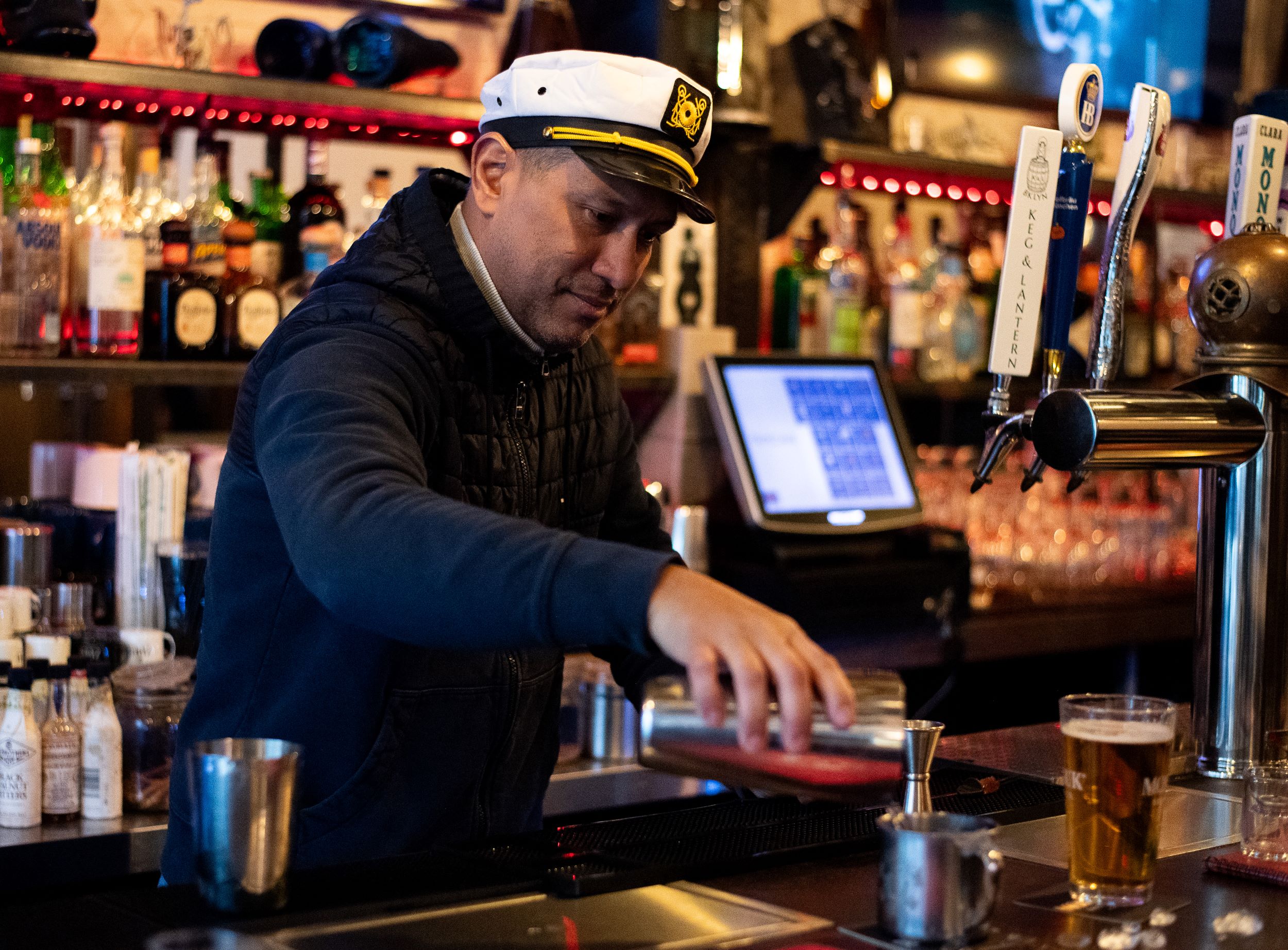 A bartender wearing a captain's hat serves drinks at a bar, with beer taps and various bottles of liquor in the background.