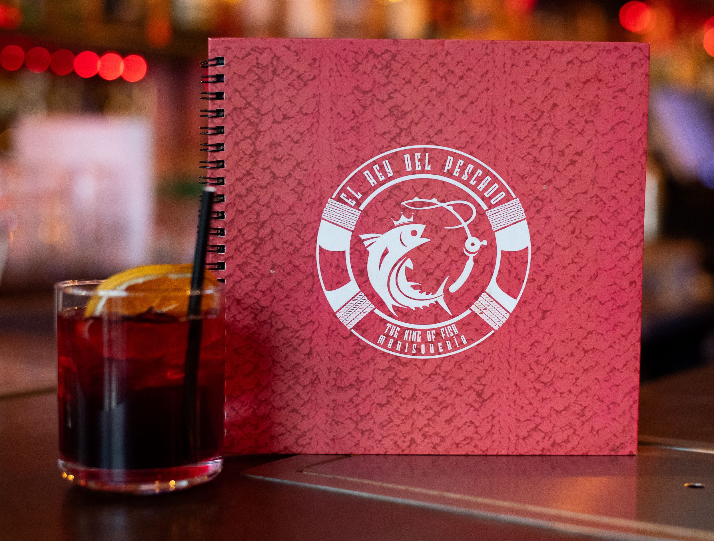 A pink, textured menu with a white and red decorative logo, next to a glass of iced drink garnished with a lemon slice, placed on a wooden bar surface.