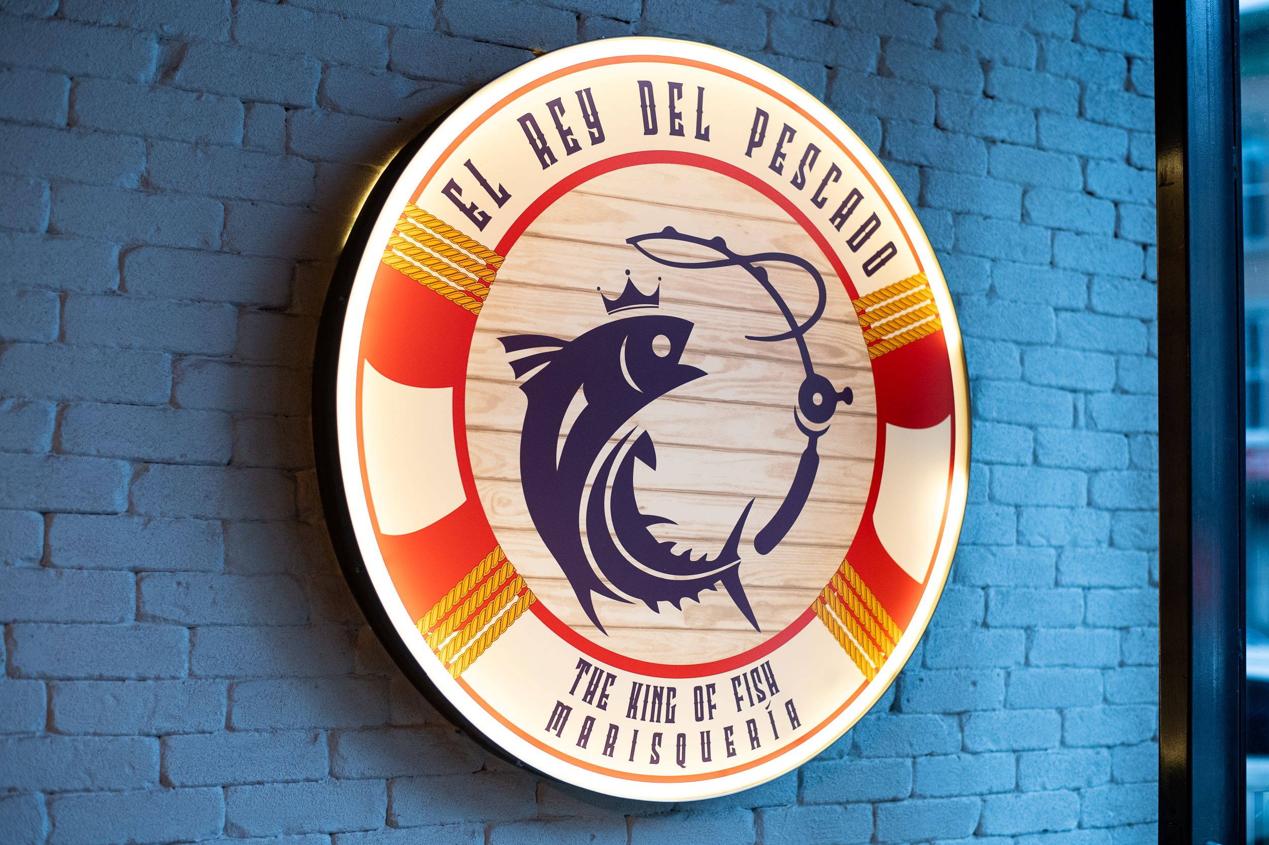 A colorful circular sign on a blue brick wall reading "el rey del pescado - the king of fish marisquería," featuring stylized graphics of two fish and sun rays.
