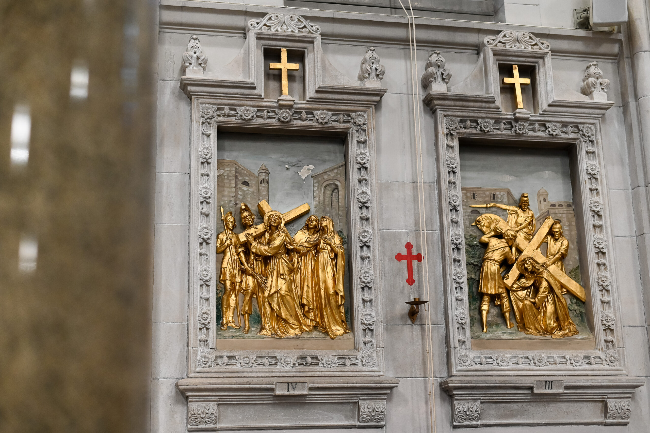 Religious carvings in gold depicting biblical scenes on church doors, flanked by crosses, in an architectural setting with a glimpse of a reflective surface on the left.