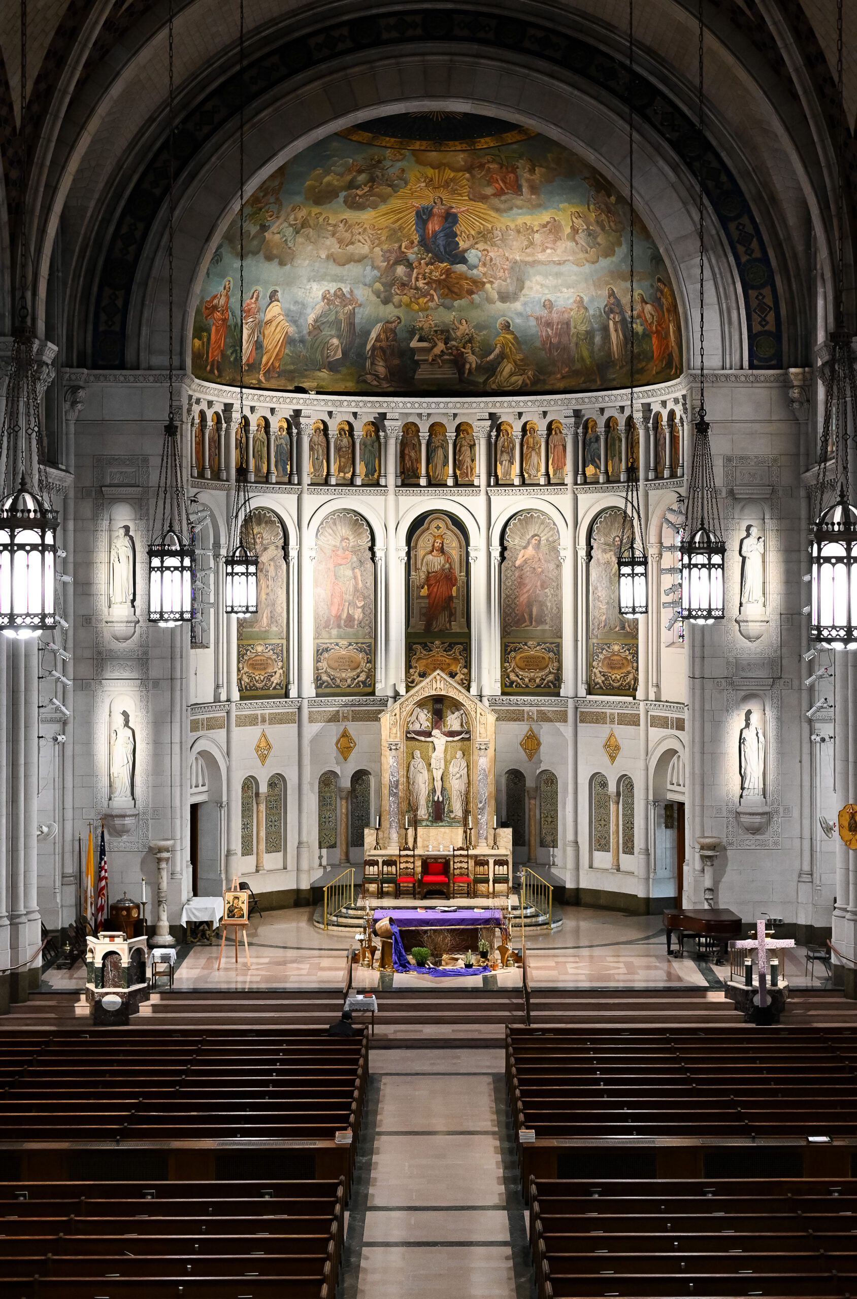 Interior of a cathedral featuring a large altar, ornate religious paintings, and rows of pews viewed from the aisle. high vaulted ceilings and hanging lights add to the grandeur.