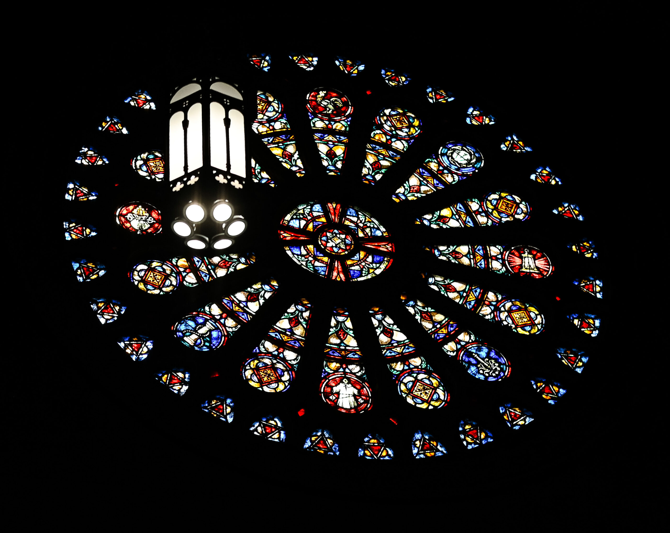A vibrant rose window in a church, featuring intricate stained glass patterns in various colors, centered by a circular clear glass pane surrounded by smaller, colorful designs.