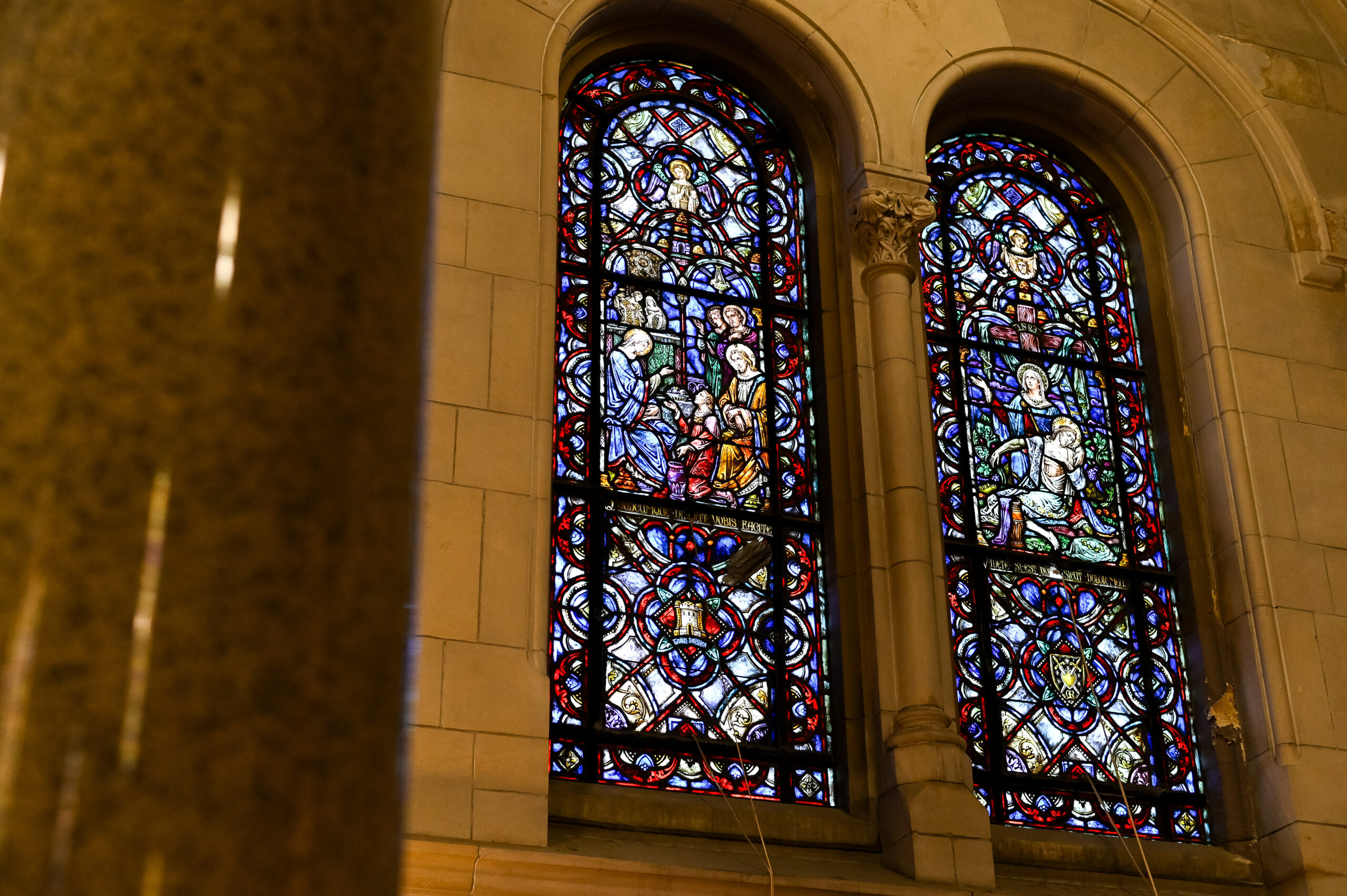 Two ornate stained glass windows in a stone building, illuminated from within, showcasing intricate designs and vibrant colors with religious imagery.