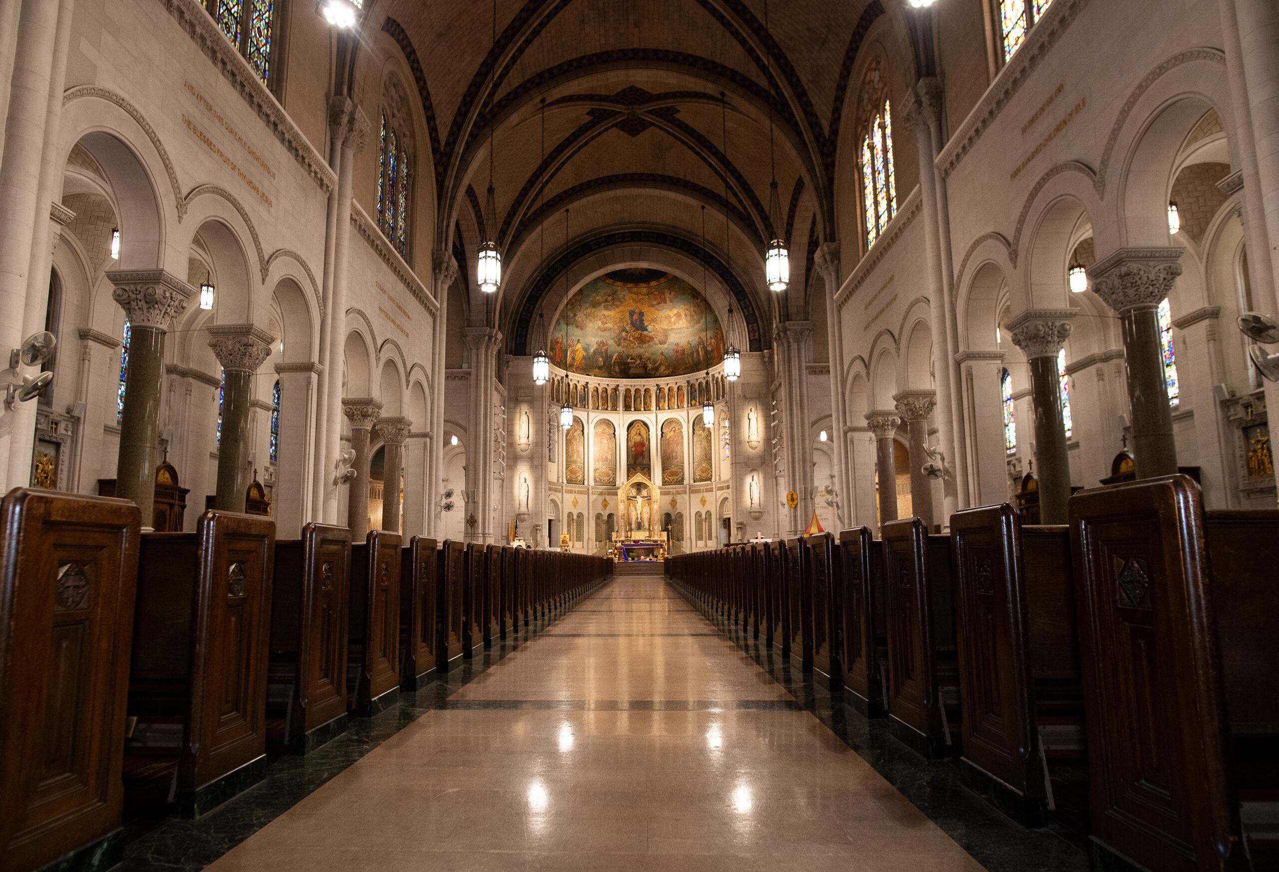 Interior of a cathedral with a long central aisle flanked by wooden pews, leading to an altar with ornate decorations and stained glass windows. arched ceilings feature detailed artwork.