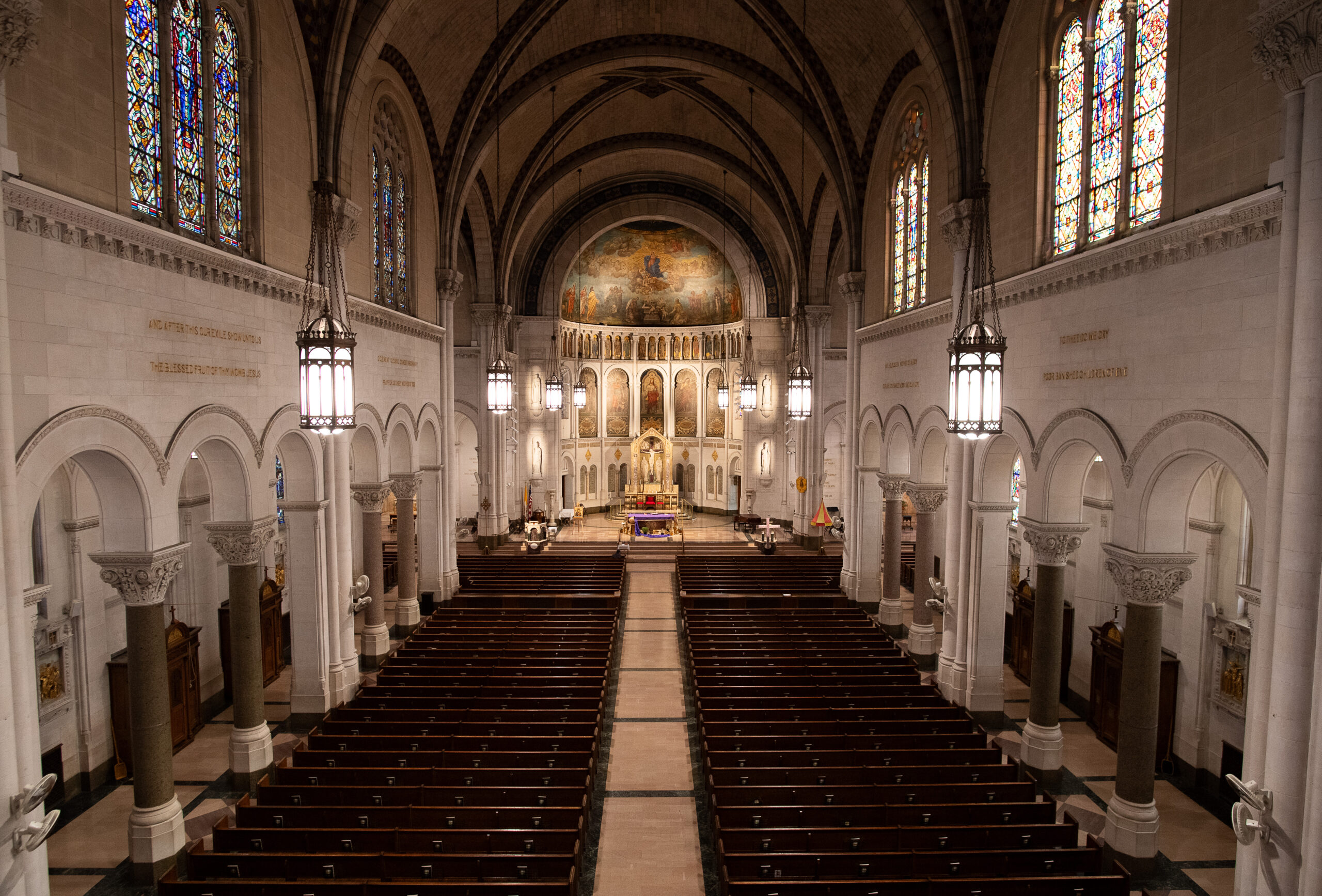 Interior view of a cathedral showcasing rows of pews leading to an ornate altar, stained glass windows, arches, and religious frescoes on the apse.