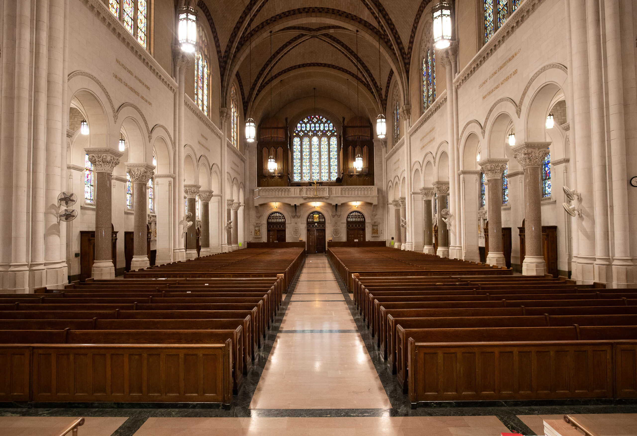 Interior of a cathedral showing long rows of wooden pews leading to an altar, with ornate stained glass windows, arched ceilings, and intricate columns.