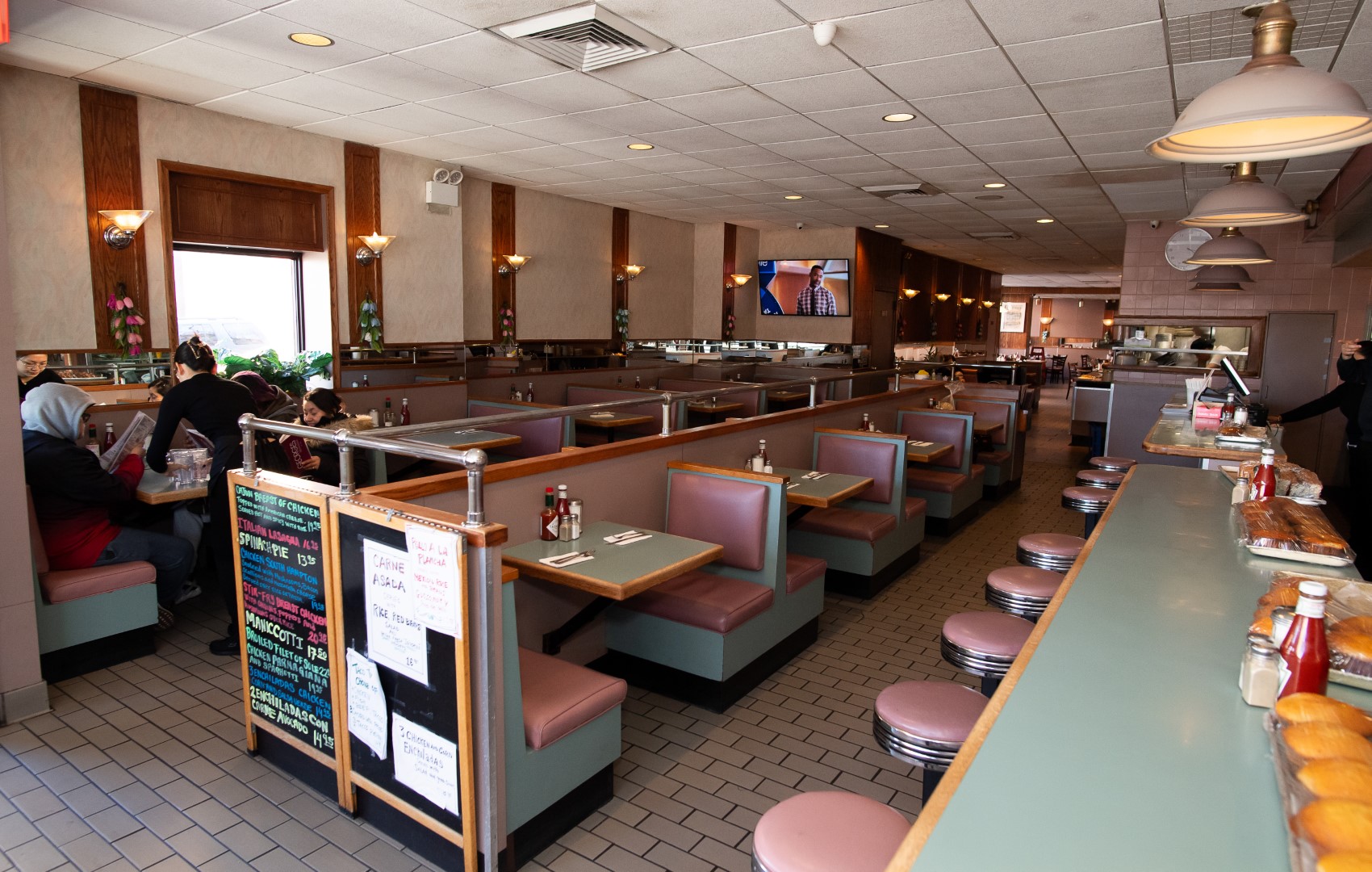 Interior of a bustling diner with pink and green booths, a counter with stools, a display of baked goods, and customers dining. a tv screens sports in the background.