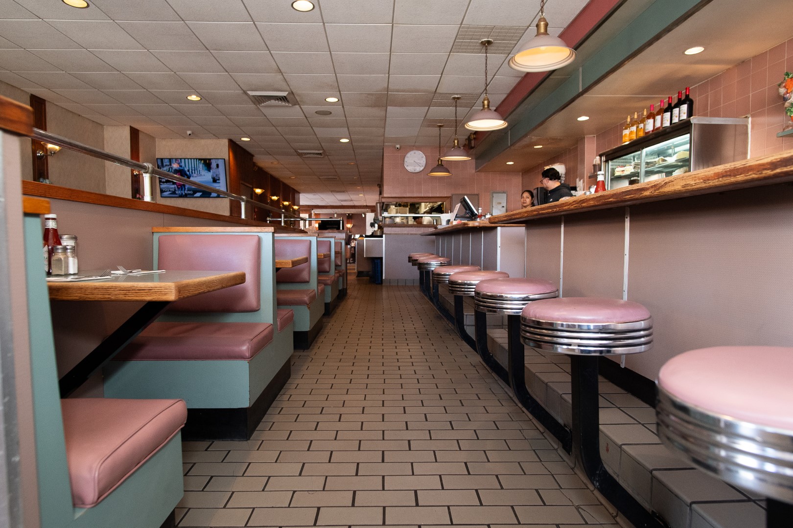 Interior of a classic diner featuring rows of pink and teal booths, chrome bar stools, and a tiled floor, with a counter and staff visible in the background.