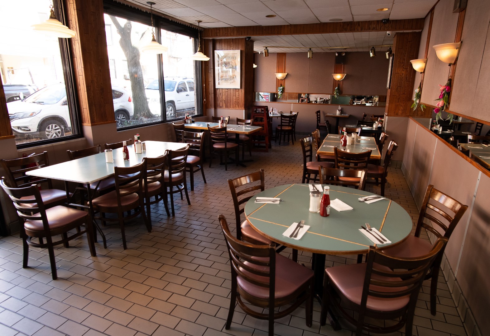Interior of a casual dining restaurant with multiple empty tables and chairs, tiled floor, and large windows with street view. each table is set with condiments and napkins.