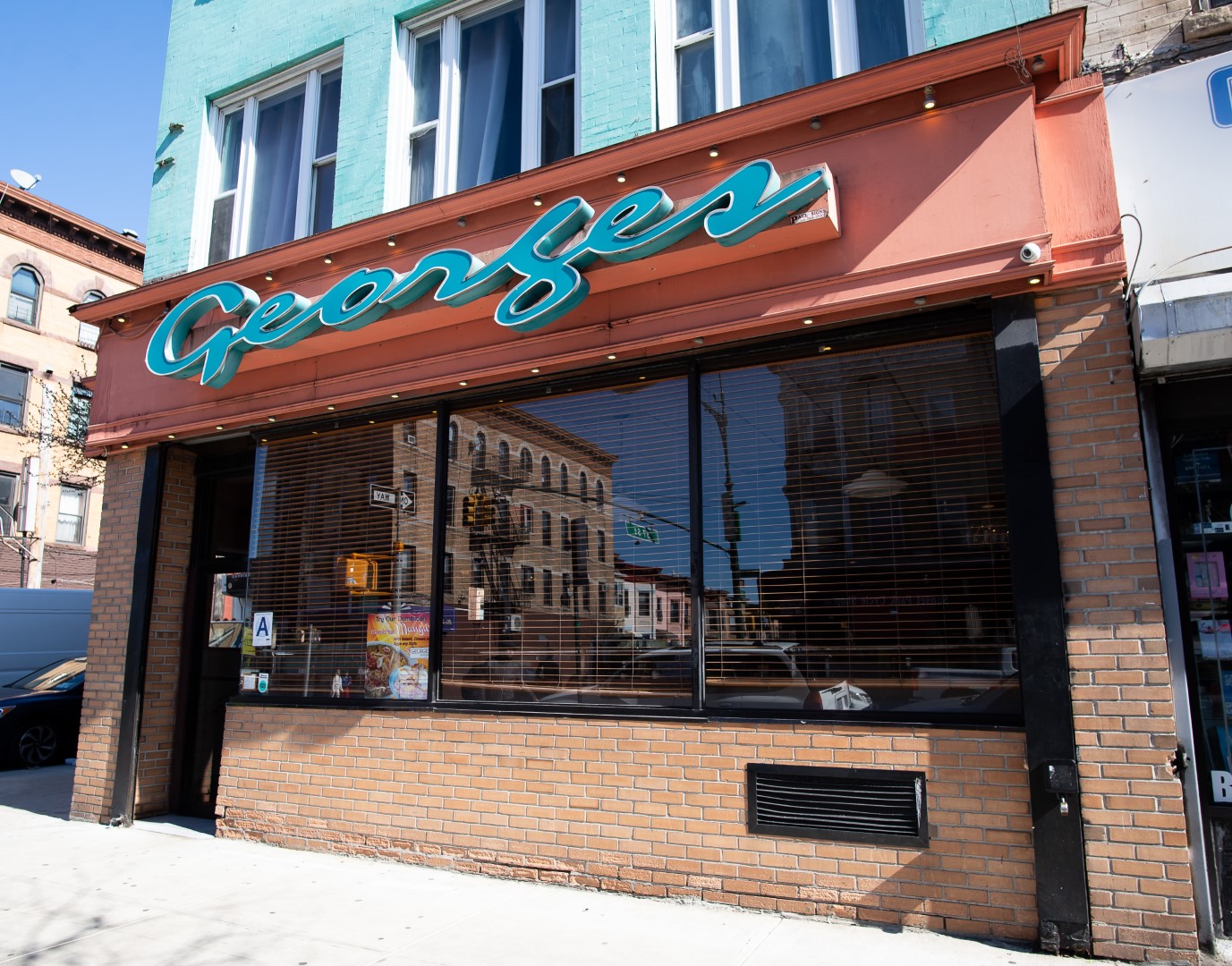 Exterior view of george's, a restaurant with a teal neon sign above large windowed facade reflecting the street, set in a brick building with a blue-green upper section.