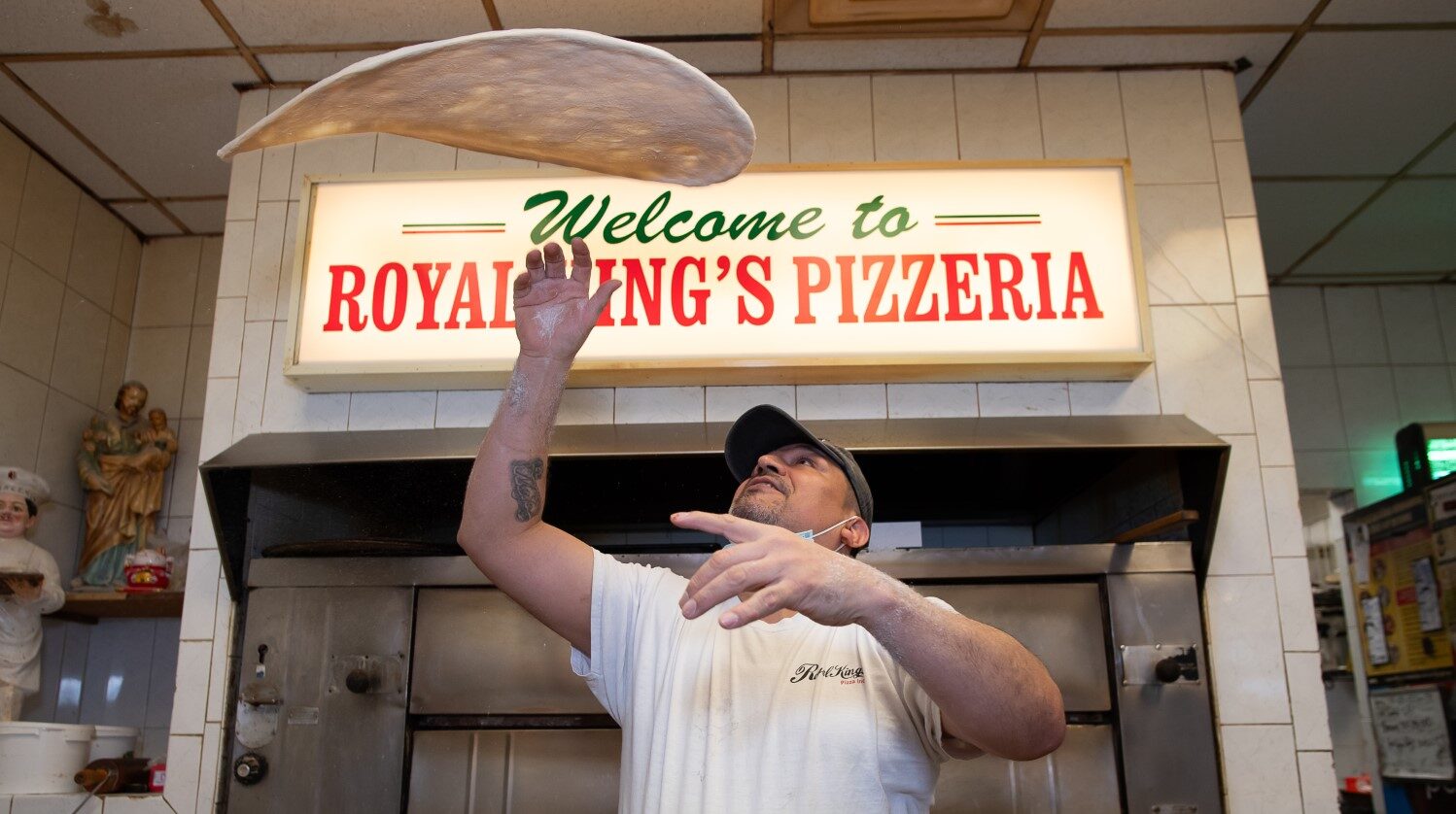 A pizza chef expertly tosses dough in the air at royal king’s pizzeria, under a welcoming sign, with decor including figurines on display.