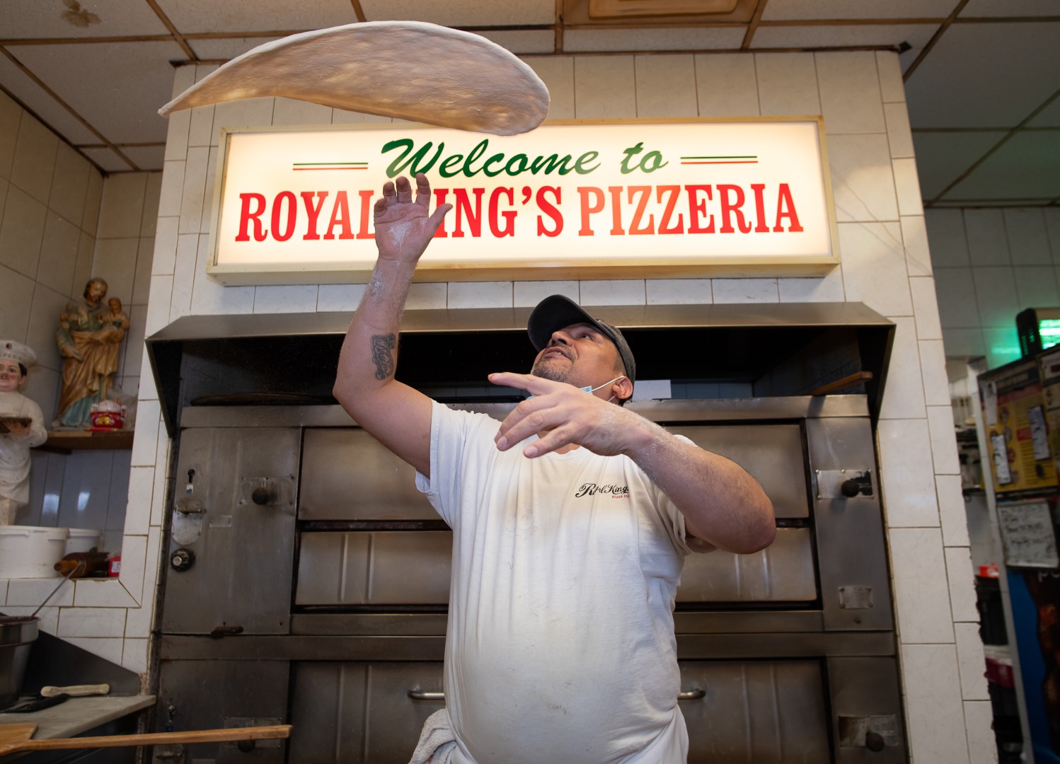 A pizzeria worker, wearing a white t-shirt and cap, skillfully tosses a stretch of pizza dough in the air inside a kitchen with a sign reading "welcome to royal king's pizzeria.