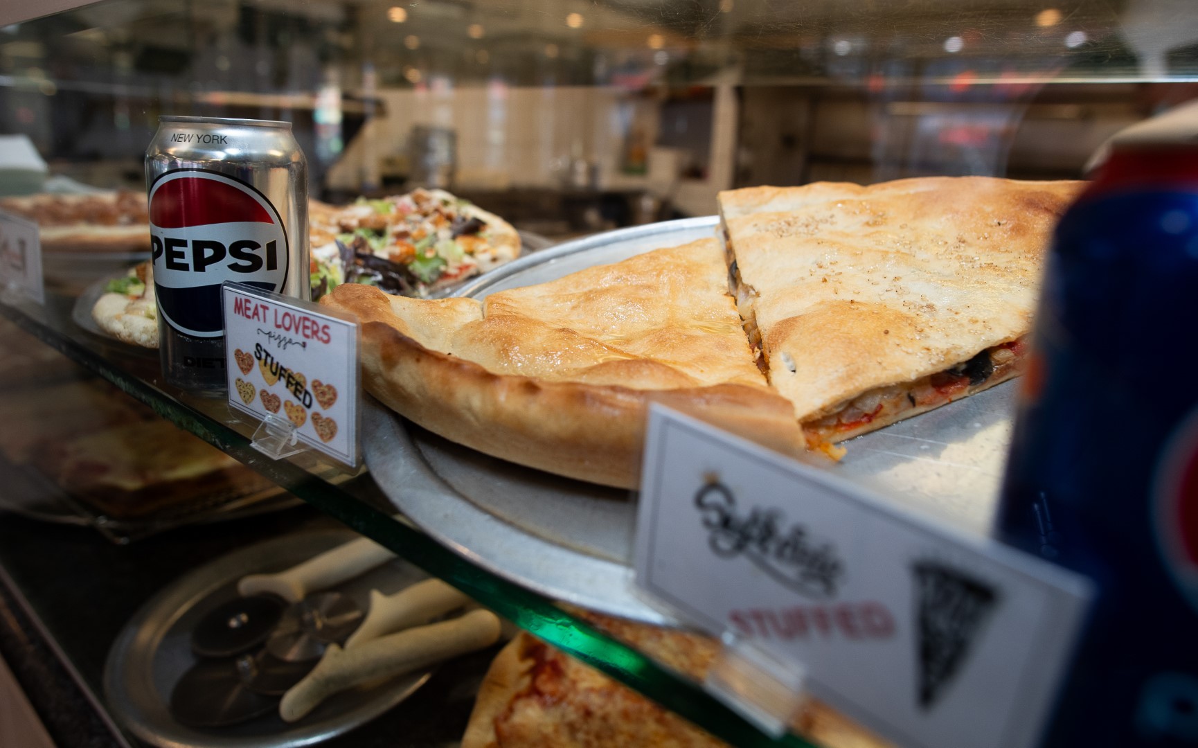 A display case at a restaurant featuring a variety of pizzas, including a "meat lovers" and a "stuffed" pizza, with a can of pepsi visible in the background.