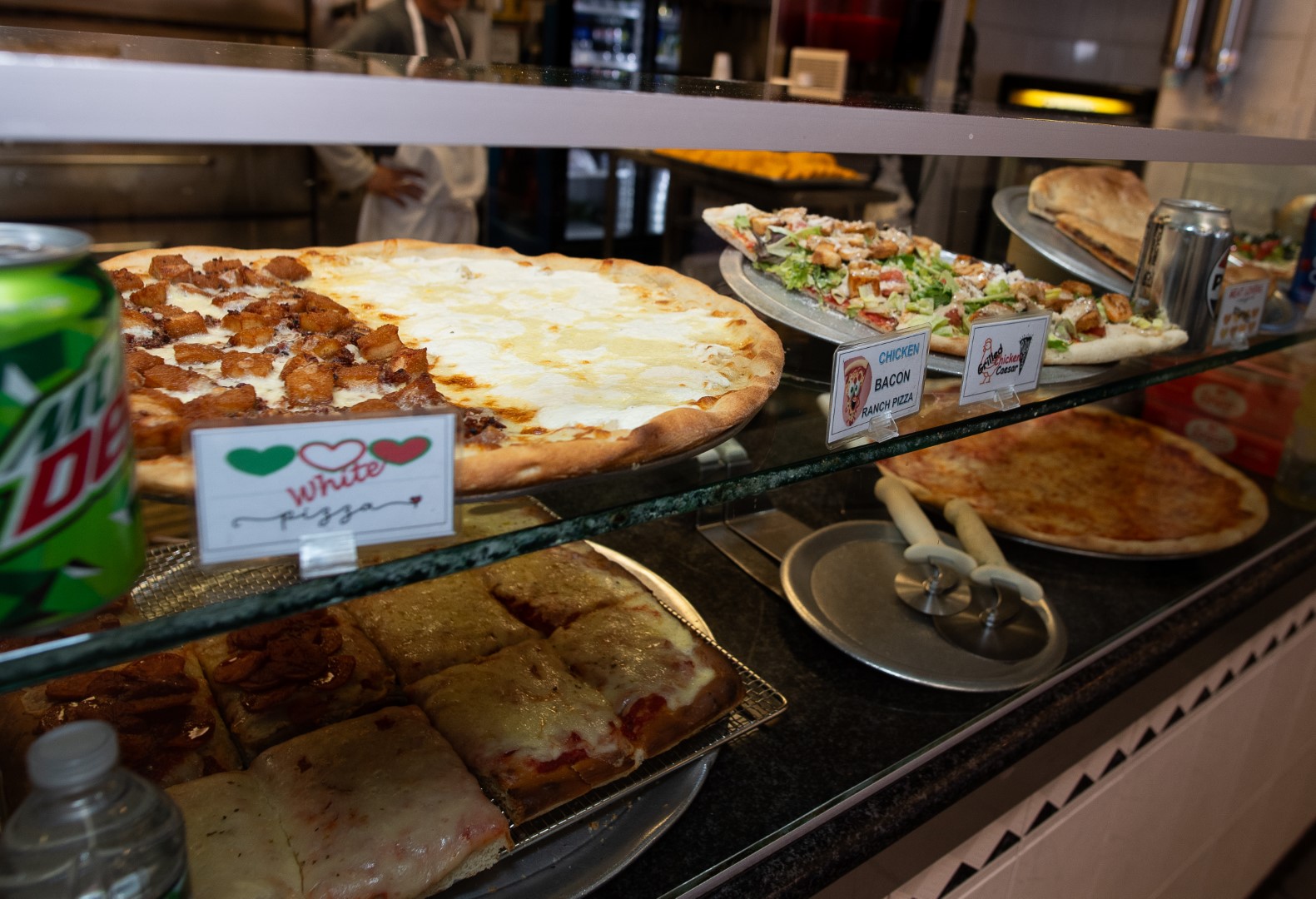 Display of various pizza slices in a pizza shop, with labels like "white pizza" and "bacon chicken ranch," visible through a glass counter, with a soda can on the side.