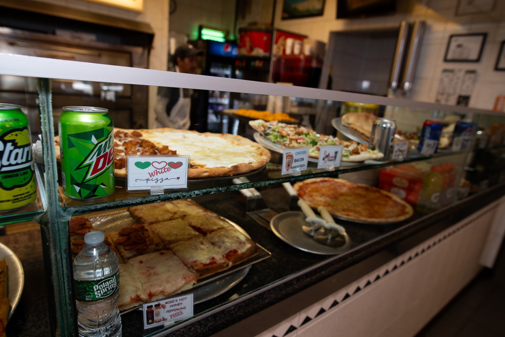 A display case at a pizzeria showcasing various pizza slices, snacks, and beverages. visible are cheese pizza, veggie pizza, and lasagna. staff are seen in the background.