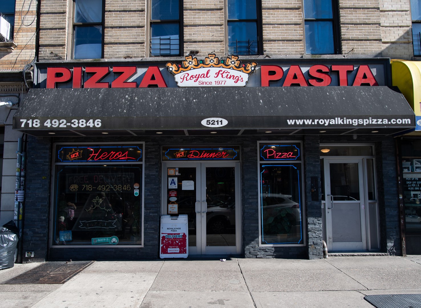 A street view of royal kings pizza pasta restaurant with signs displaying the business name, food options, phone number, and website link on a sunny day.